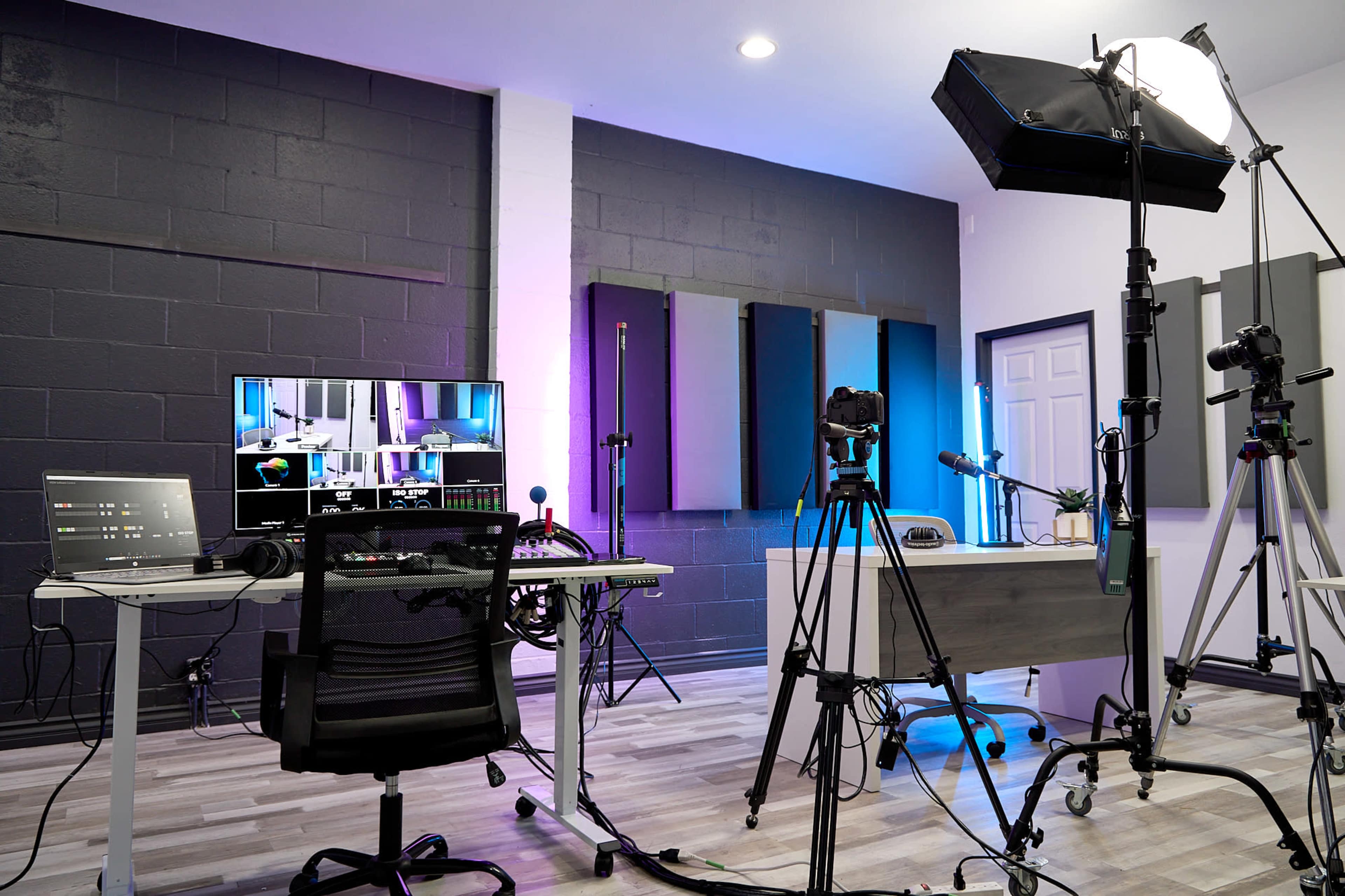 A modern recording studio setup featuring a desk with computer monitors, cameras on tripods, and soundproofing panels on the walls.