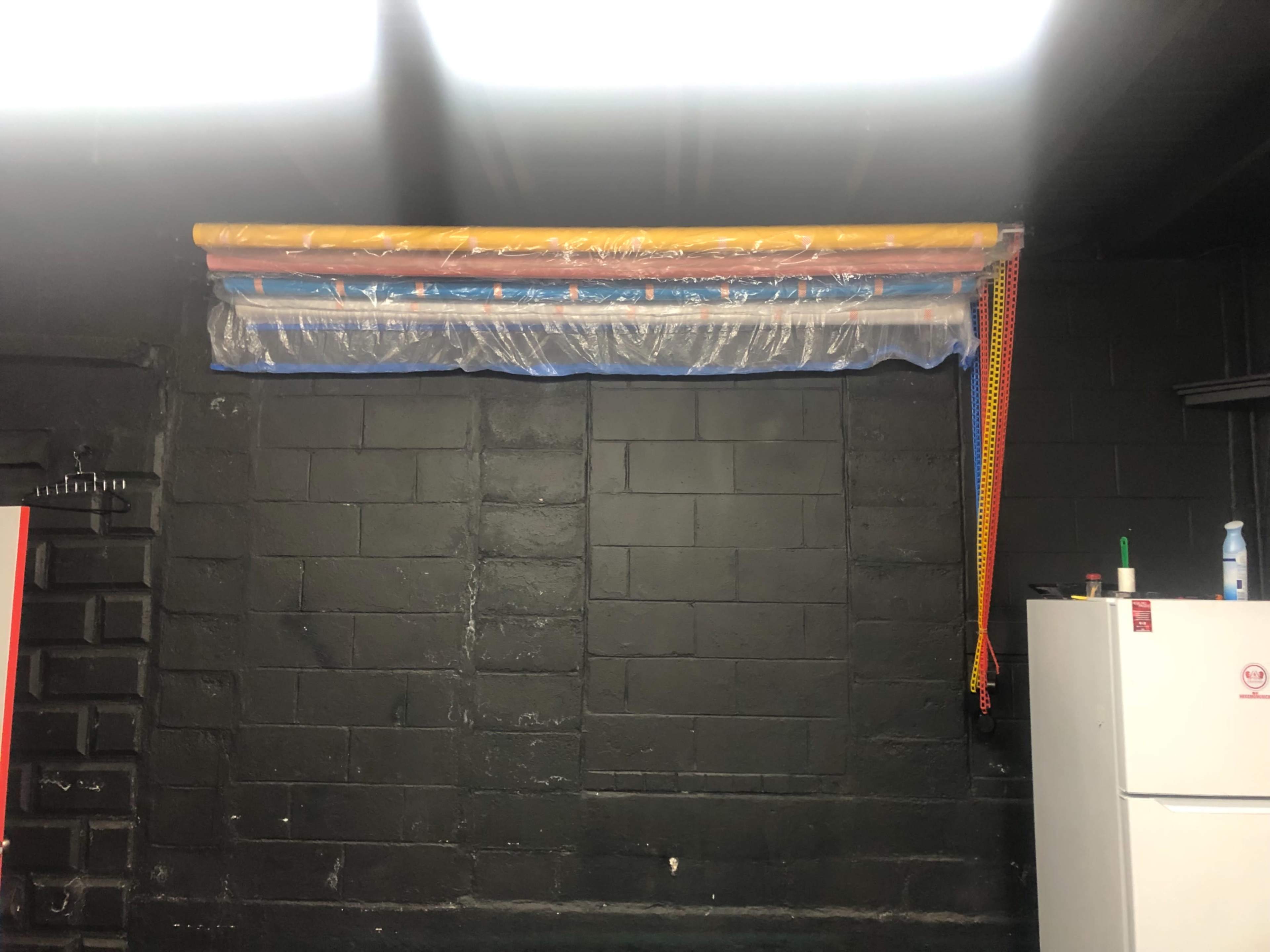 The image shows a wall with several long, colorful plastic tubes secured above it, alongside a white refrigerator in a dark room.
