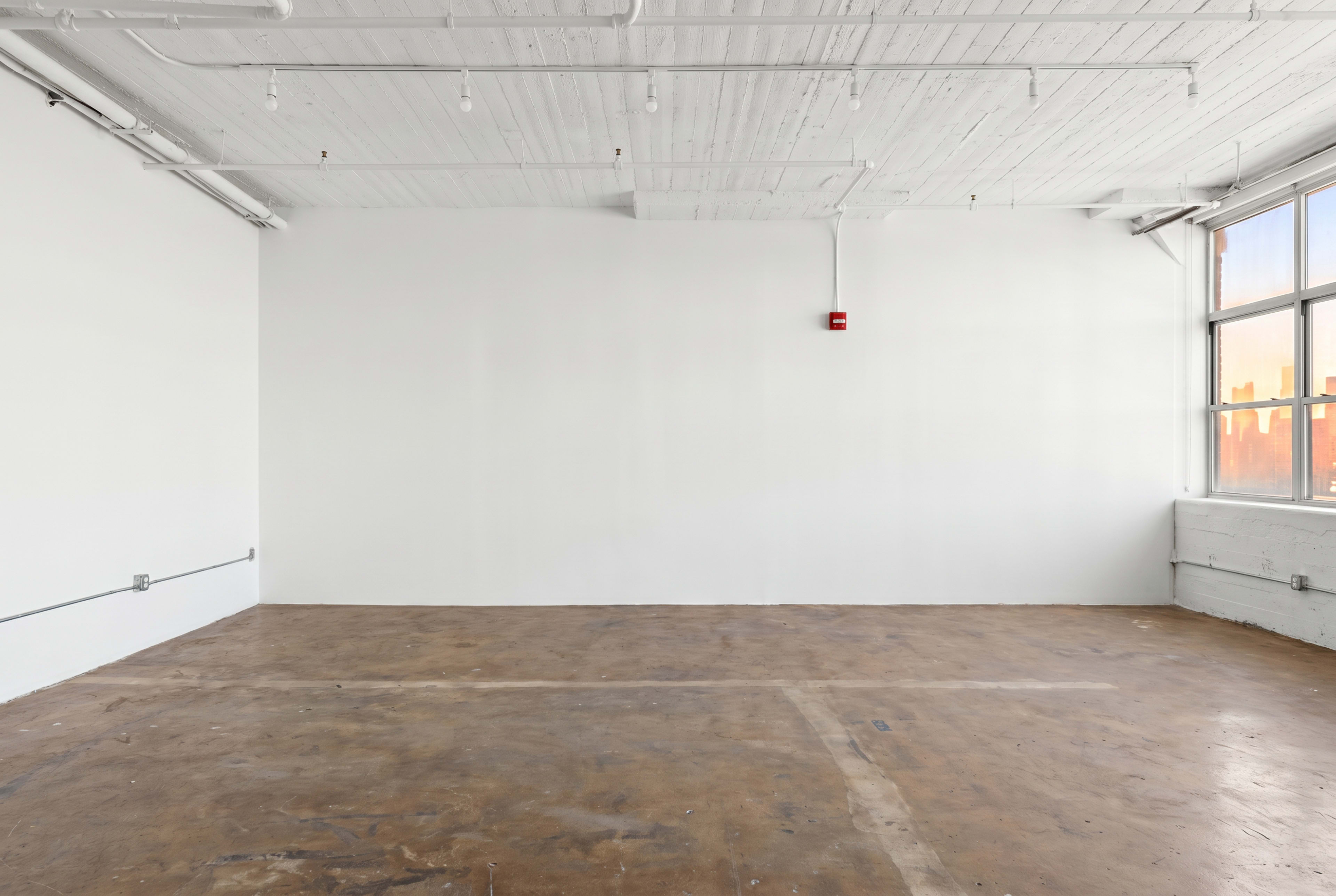 The image shows an empty room with a bare concrete floor and white walls, illuminated by light from a large window.