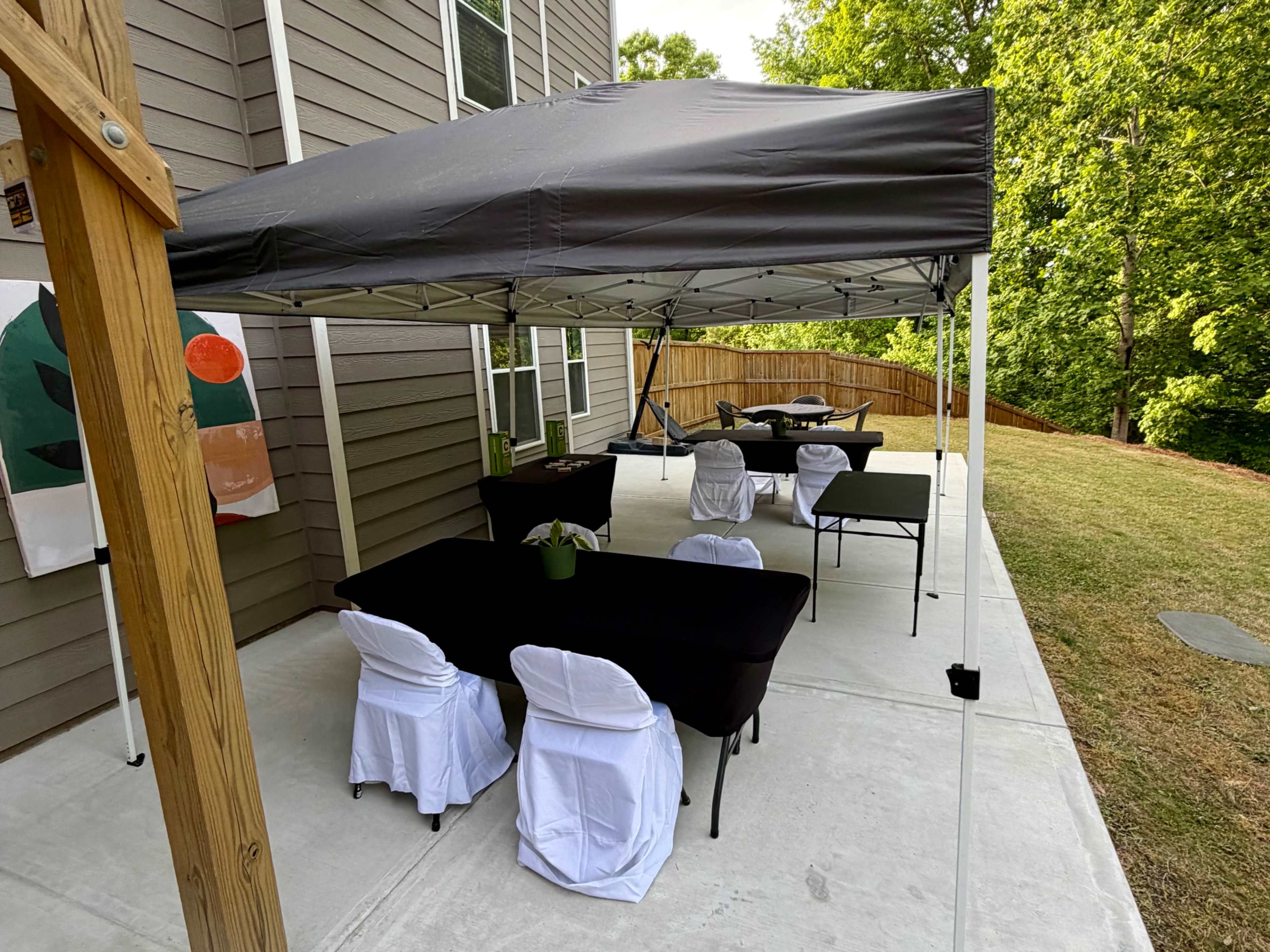 A black canopy is set up over several tables and chairs on a concrete patio beside a grassy area and wooden fence.