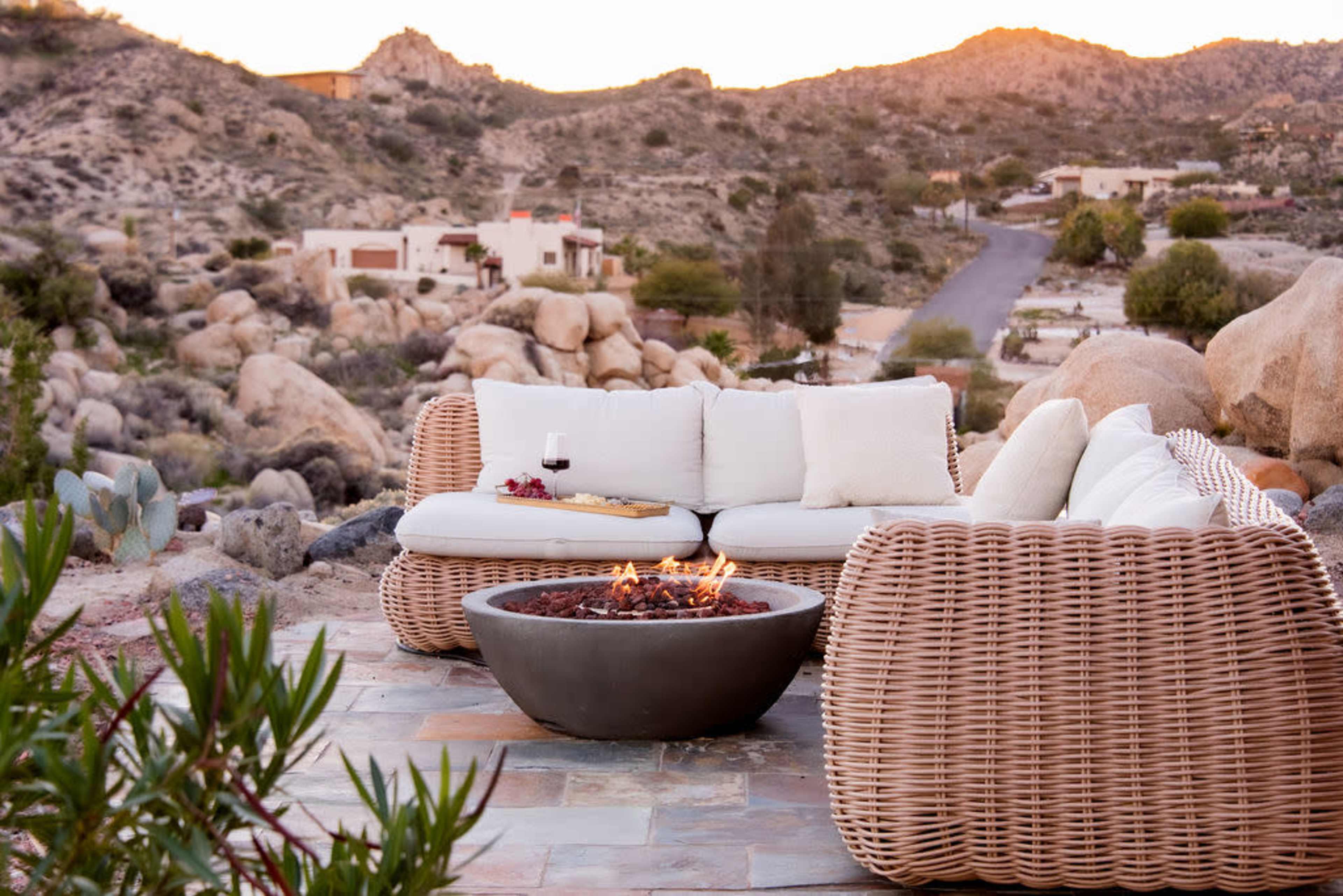 A wicker couch is arranged around a circular fire pit on a rocky patio, overlooking a desert landscape at sunset.