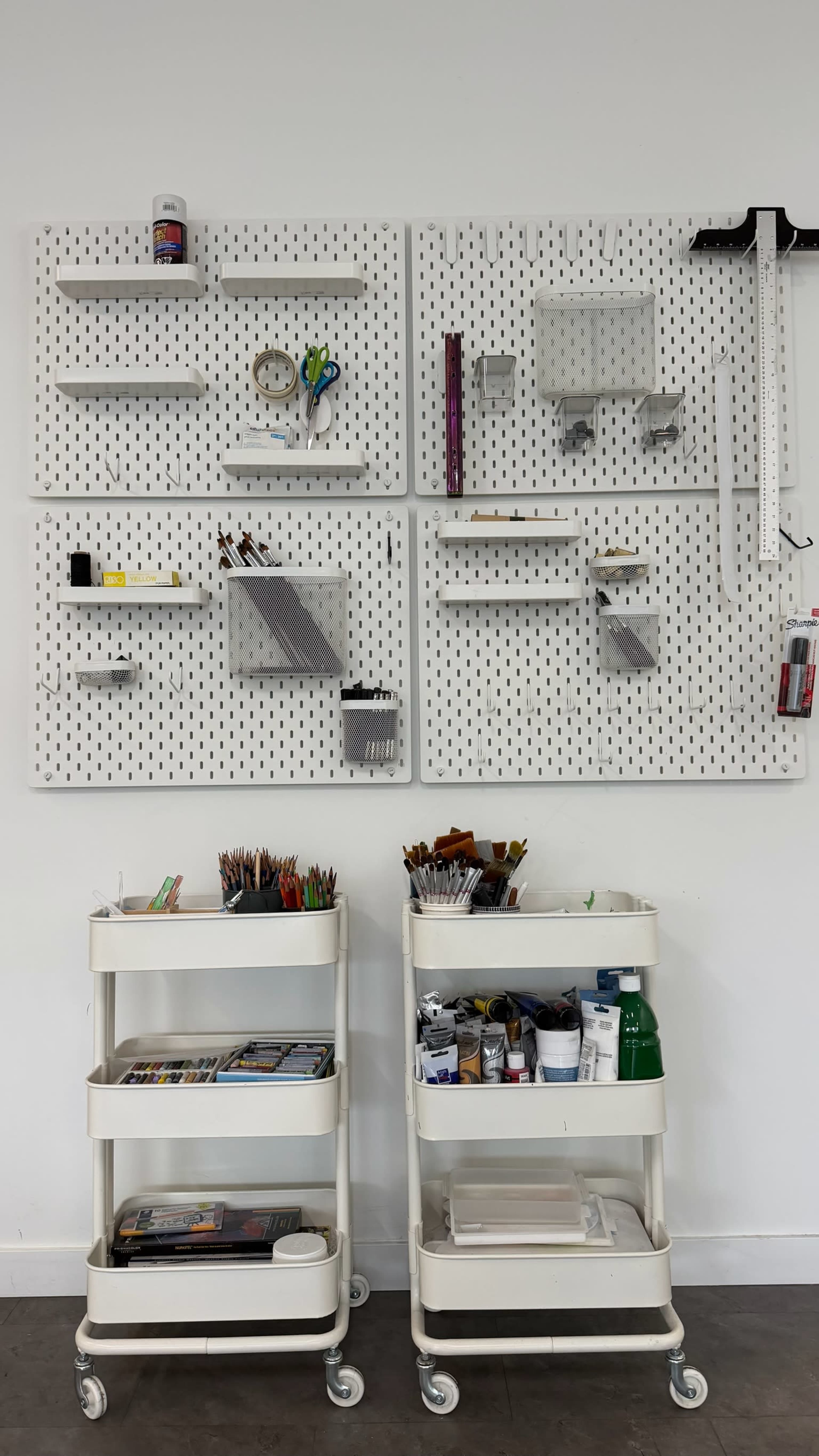 The image shows a wall-mounted pegboard display with various tools and materials organized on shelves, accompanied by two mobile carts filled with art supplies and documents.