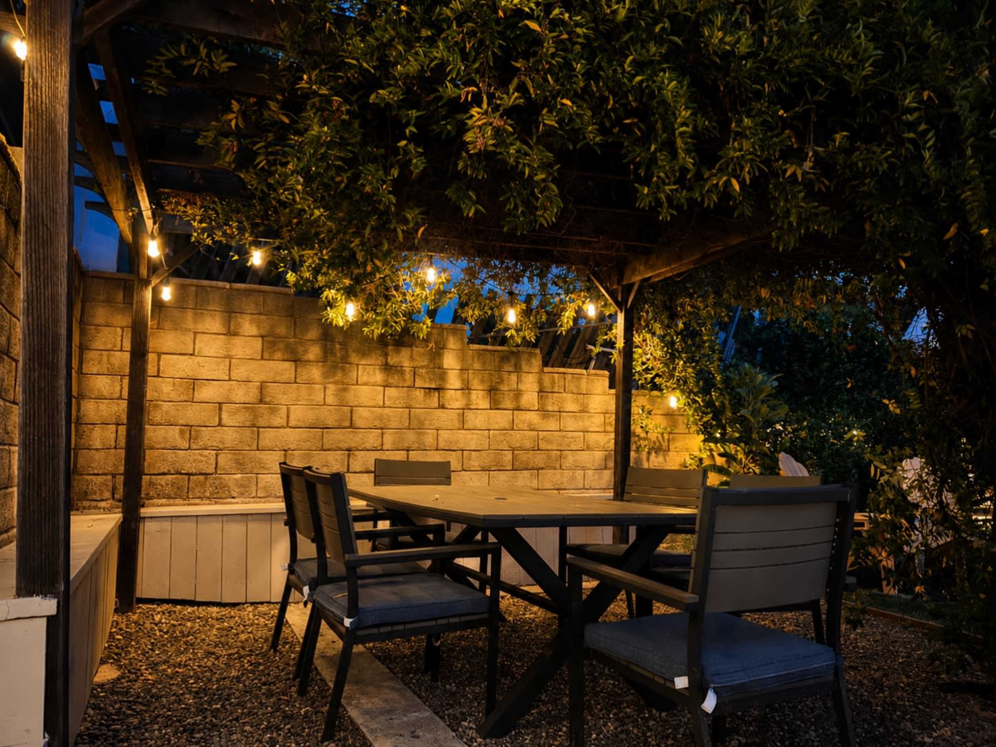 A wooden patio with a large table and several chairs is illuminated by string lights under a trellis at dusk.