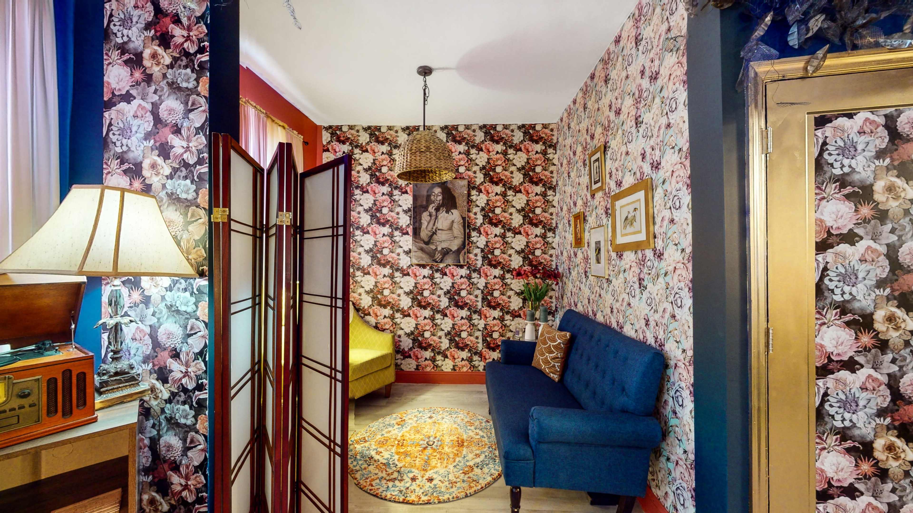 A cozy room with floral wallpaper featuring a blue sofa, a lamp, a screen divider, and a round rug, complementing a vintage radio in the corner.
