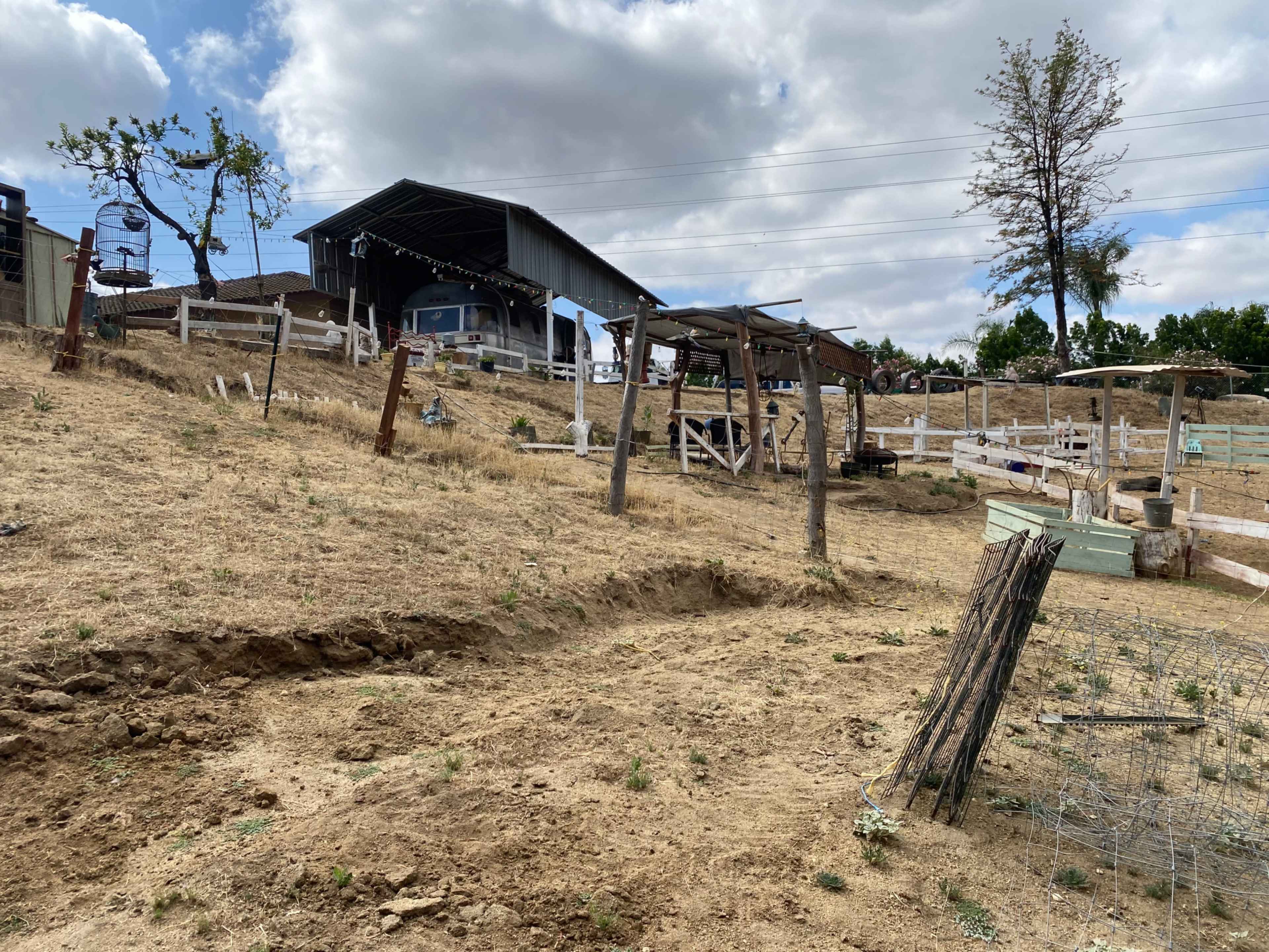 The image shows a dry, grassy hillside with wooden structures, a barn in the background, and a few trees under a cloudy sky.