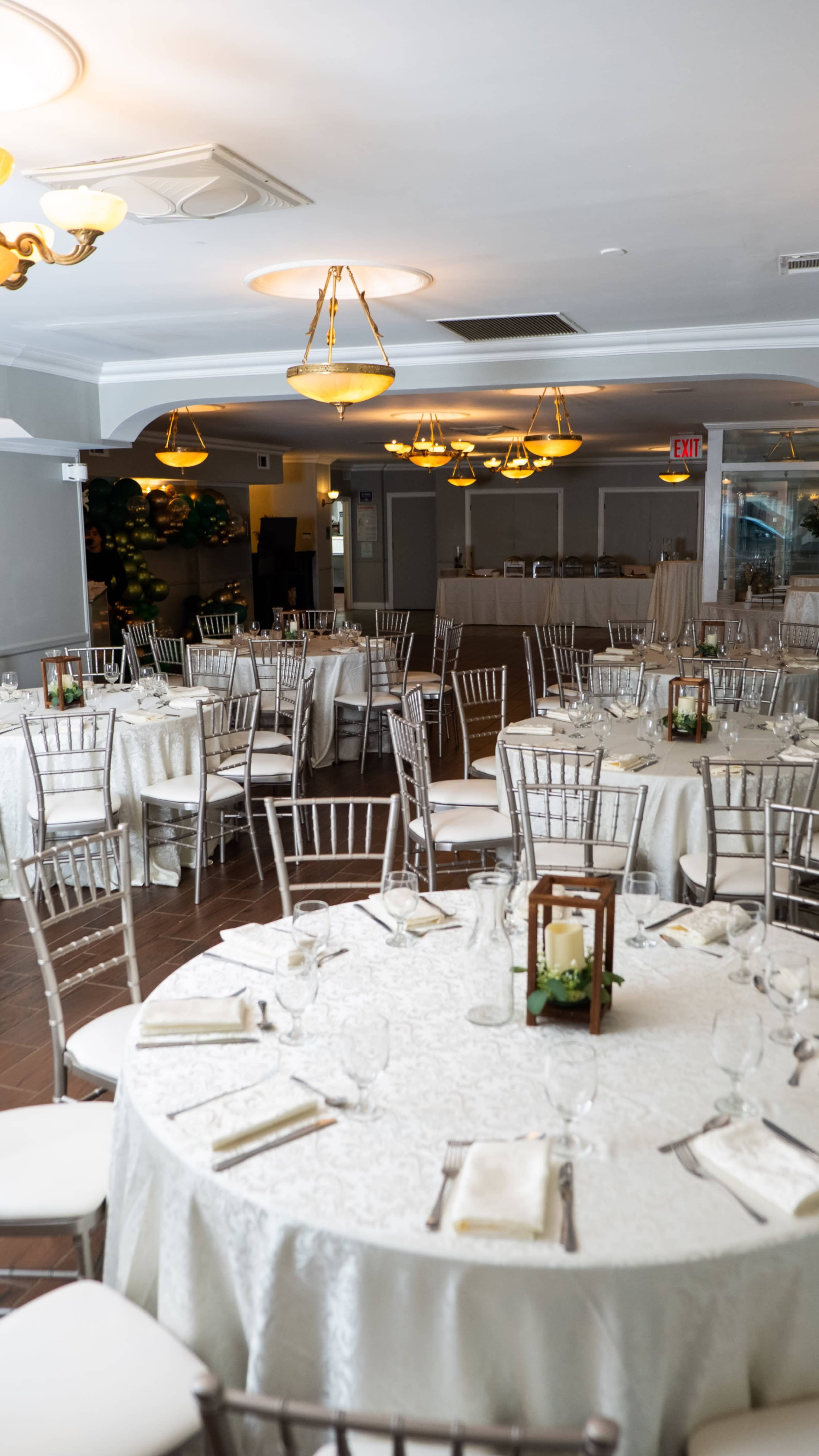 The image shows a neatly arranged dining hall with round tables covered in white tablecloths and surrounded by silver chairs.