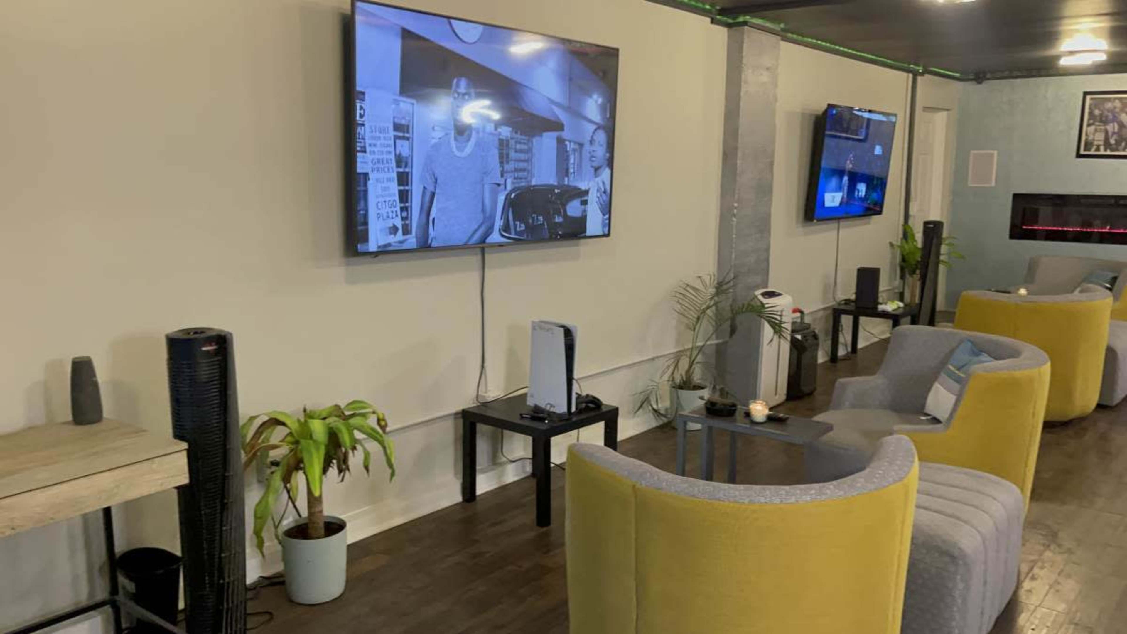 The image shows a room with a large television displaying black-and-white content, surrounded by modern seating arrangements and plants.