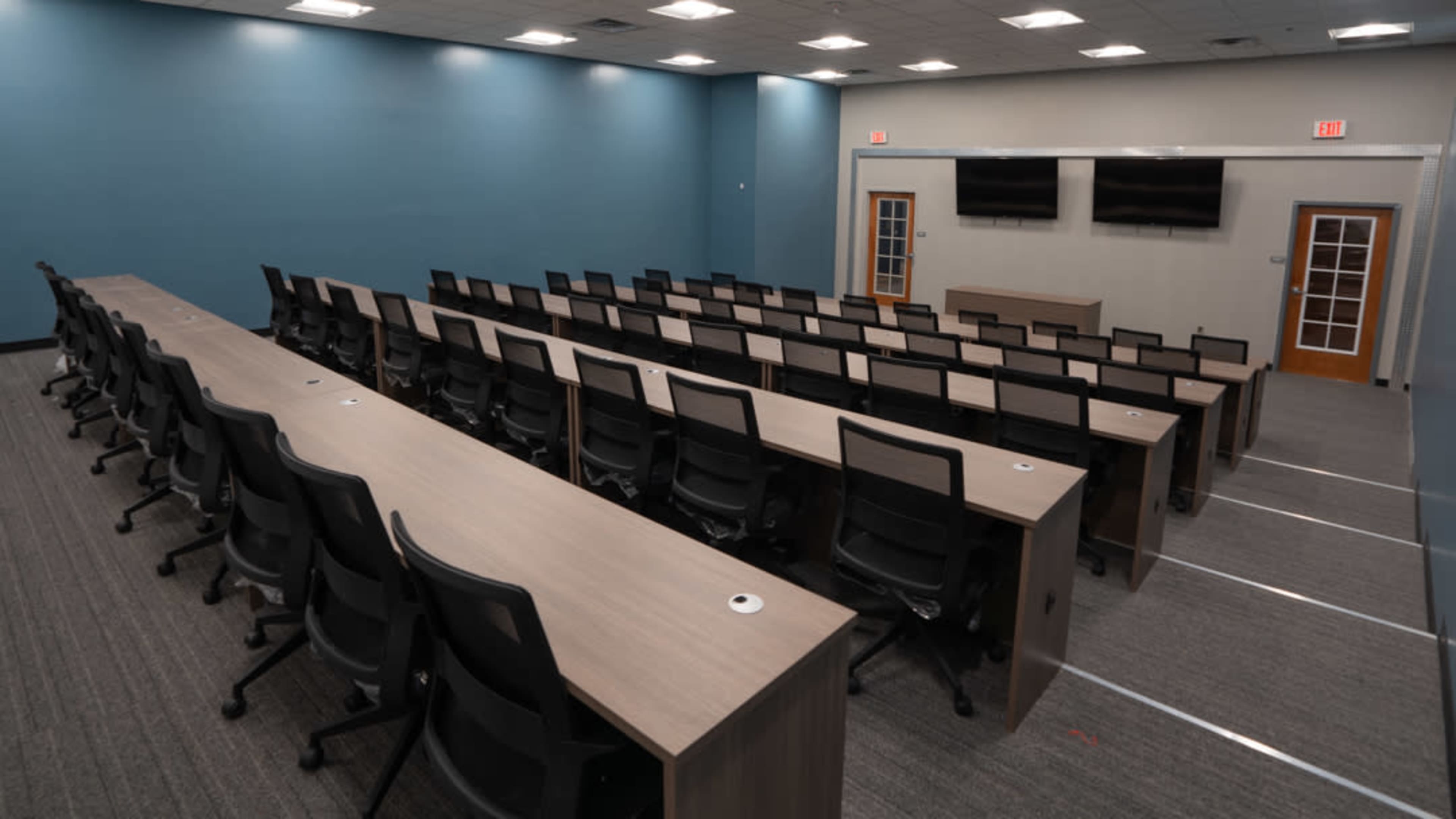 A modern classroom or conference room with rows of long tables and chairs arranged facing a presentation area, featuring two wall-mounted screens.