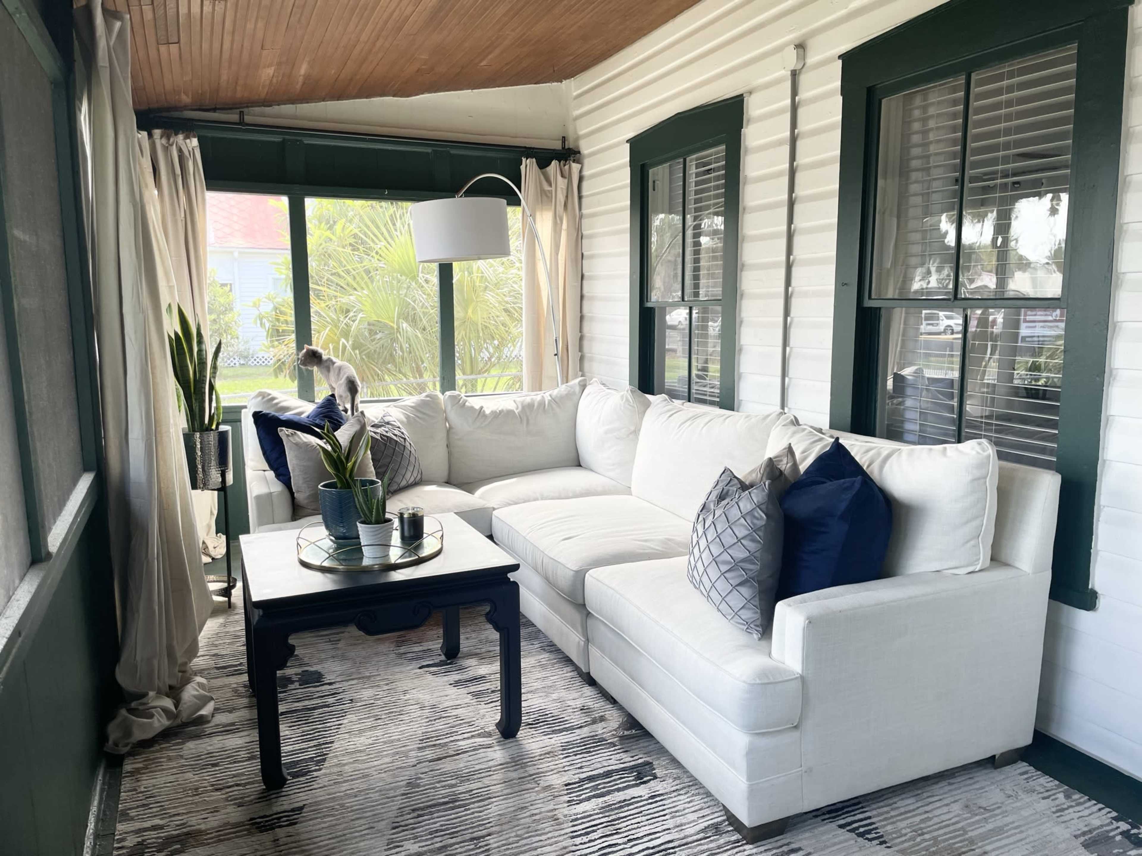 The image shows a sunlit screened porch featuring a white sectional sofa with navy and gray pillows, a dark coffee table, and potted plants, surrounded by windows and light-colored curtains.