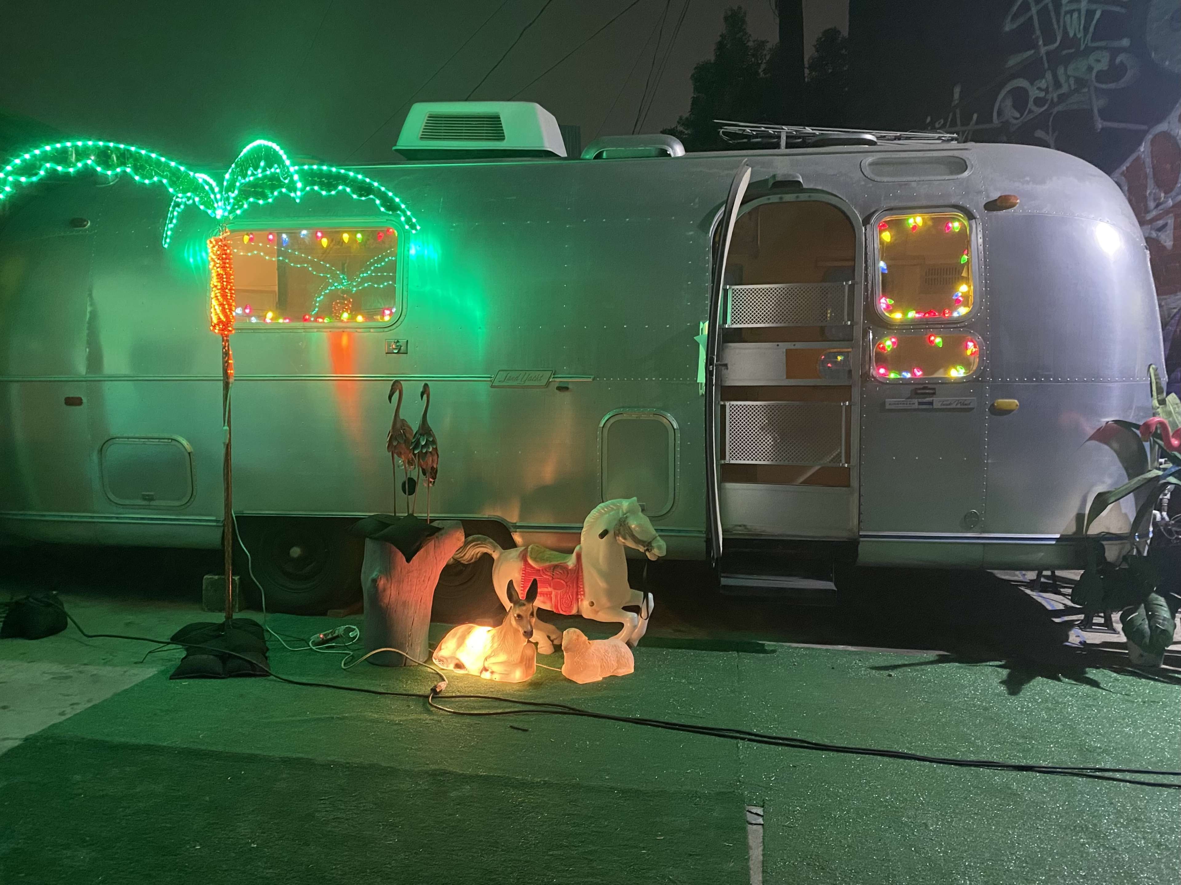 An Airstream trailer adorned with colorful lights is positioned next to illuminated decorative figures, including a palm tree and two unicorns.