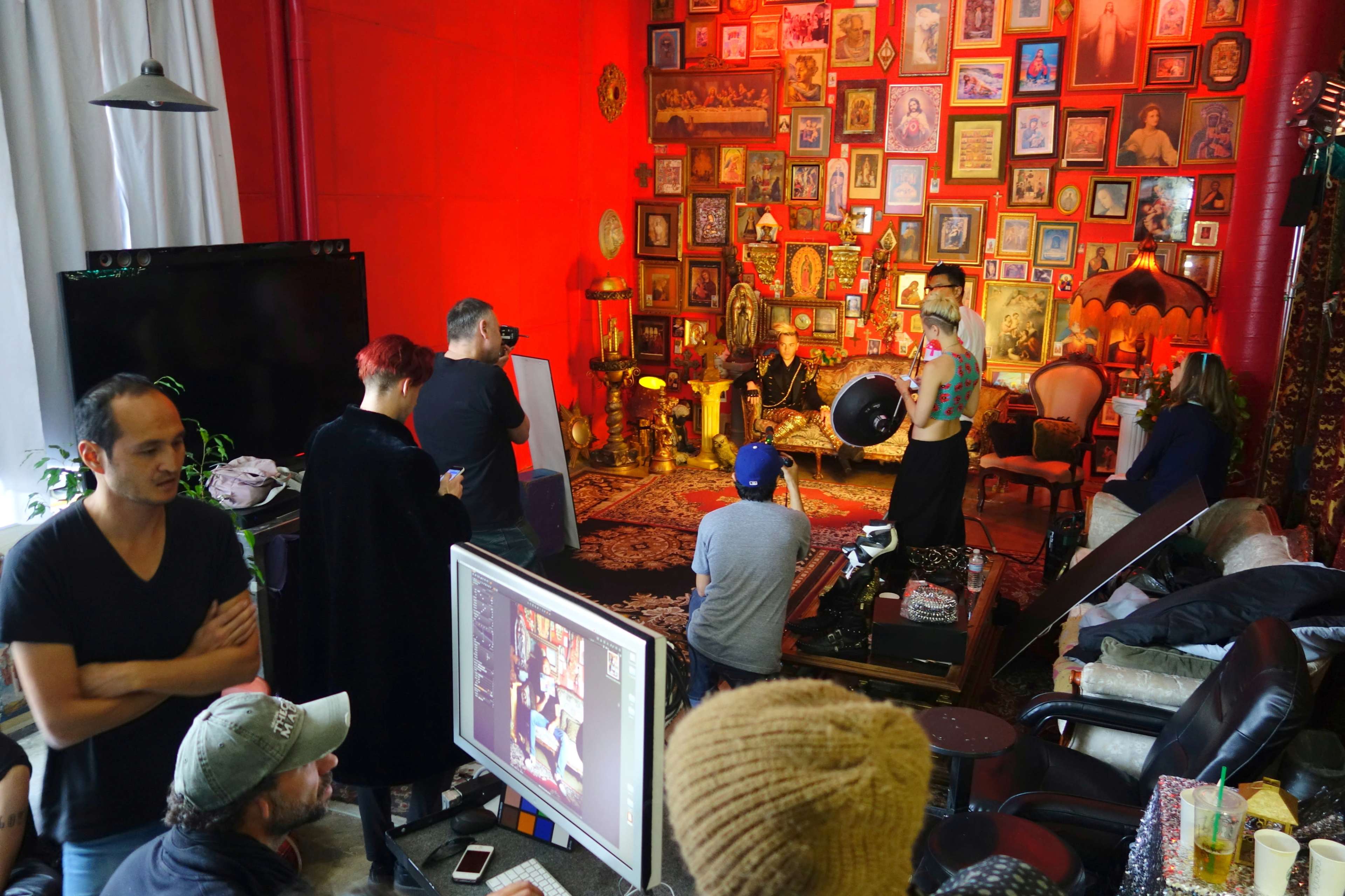 A group of people is gathered in a room with red walls, observing a photoshoot in front of a wall filled with framed pictures and decorations.