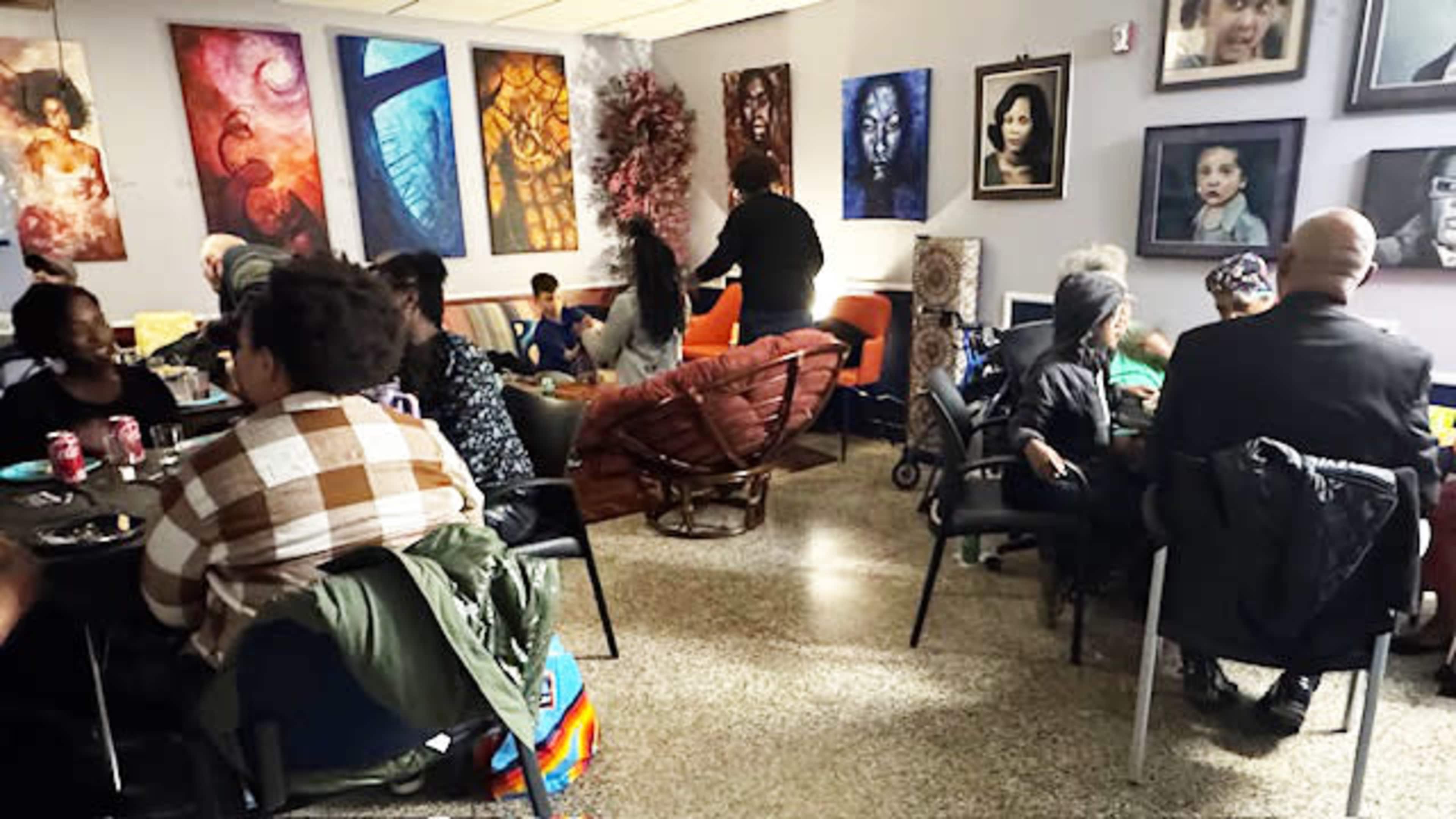 A cozy gathering space features several people seated at tables, art displayed on the walls, and a few individuals mingling in the background.