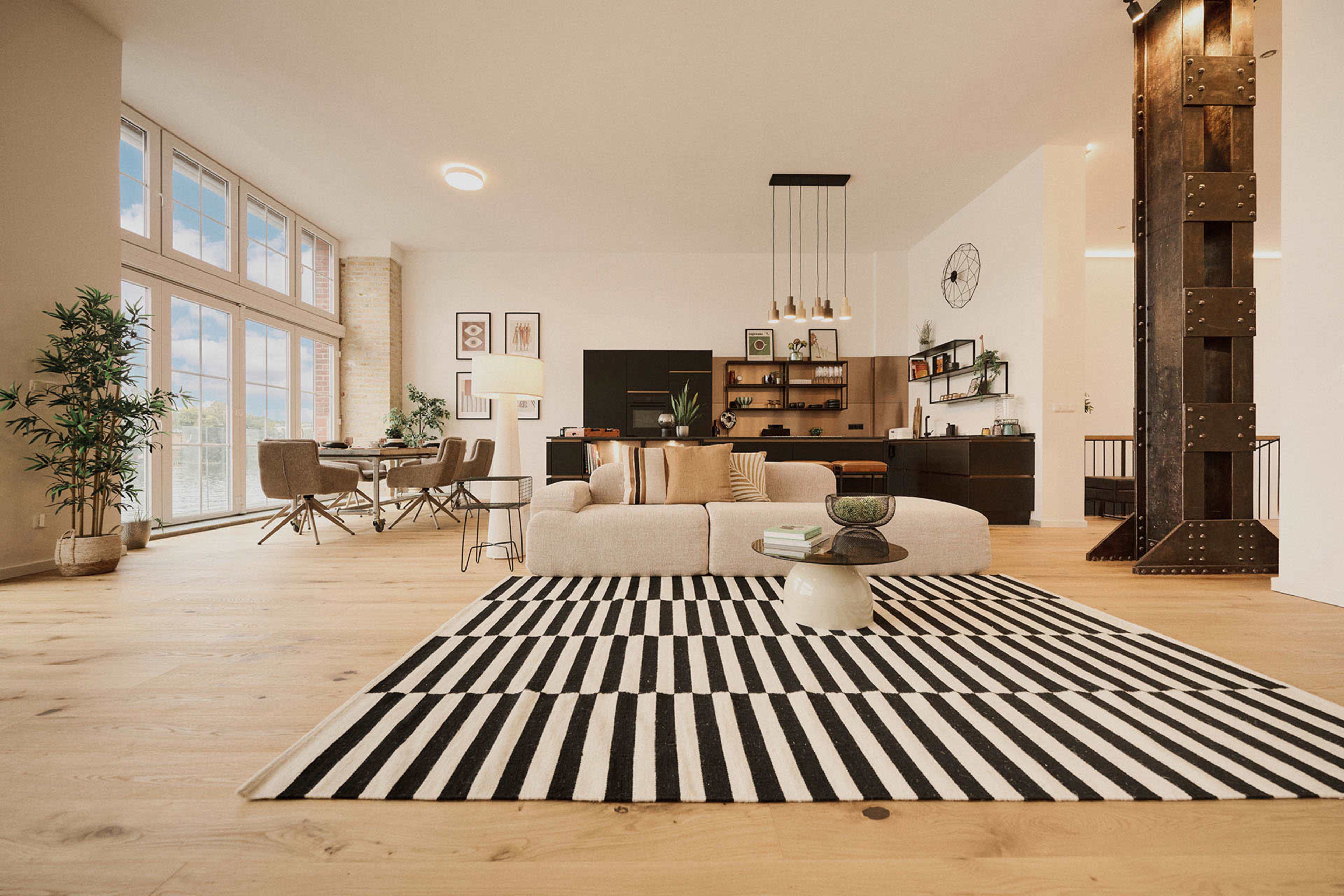A spacious living area features a light-colored couch centered on a striped rug, surrounded by modern furniture and large windows that provide ample natural light.