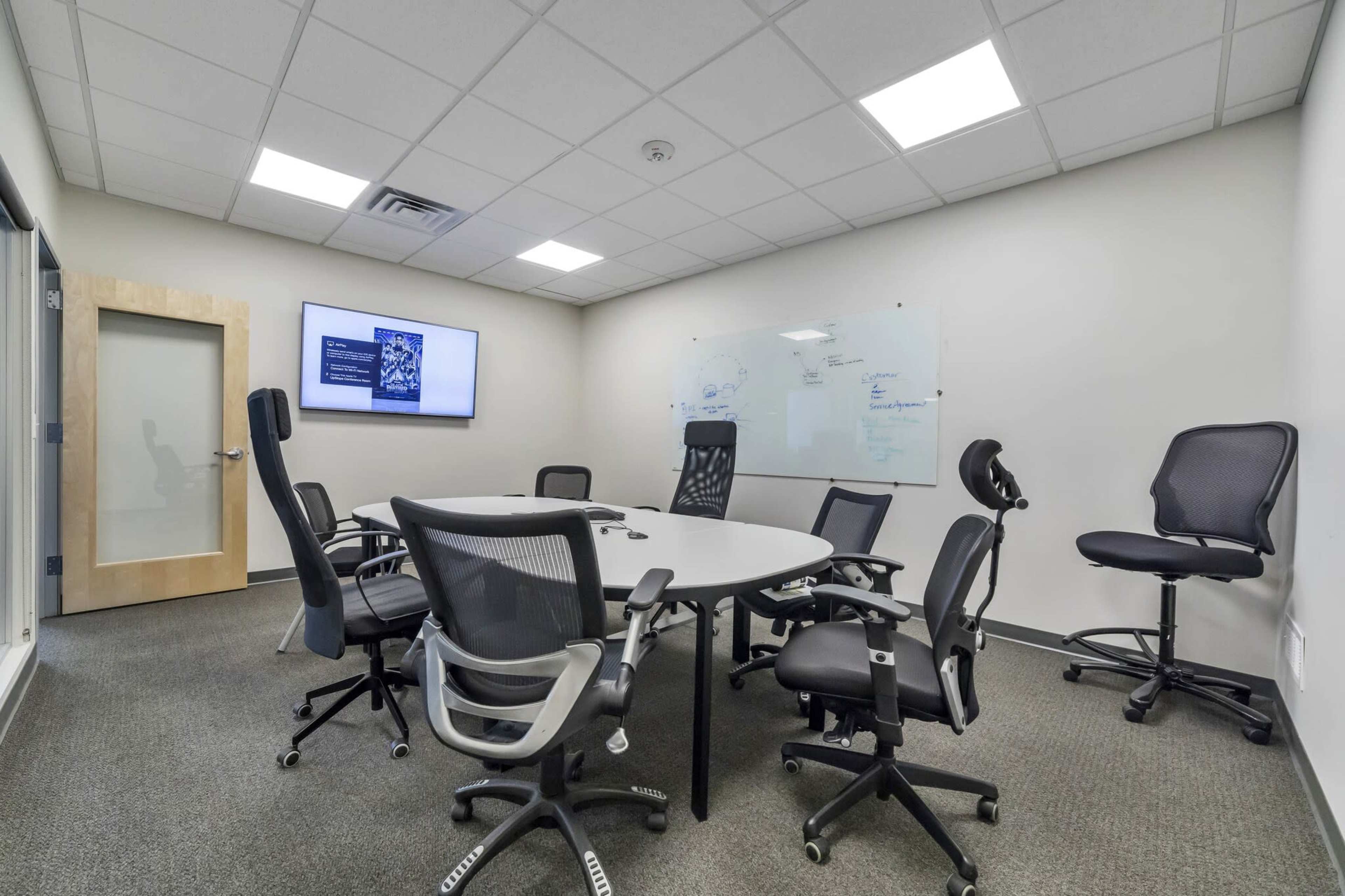 The image shows a conference room with a round table surrounded by several mesh office chairs, a glass whiteboard on one wall, and a presentation screen displaying information.