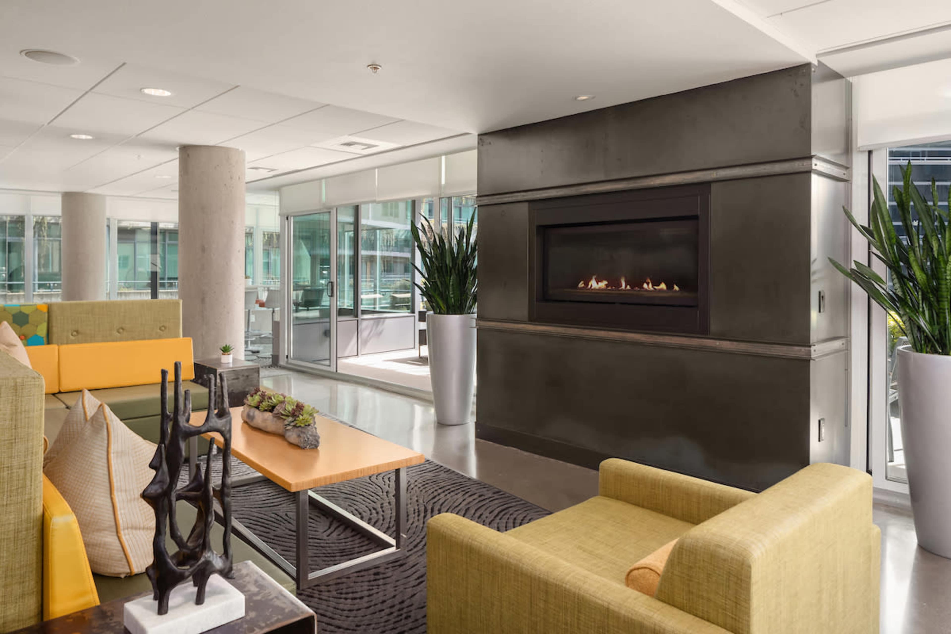 A modern lounge area features a sleek gas fireplace, green and yellow seating arrangements, and large windows that allow natural light to flood the space.