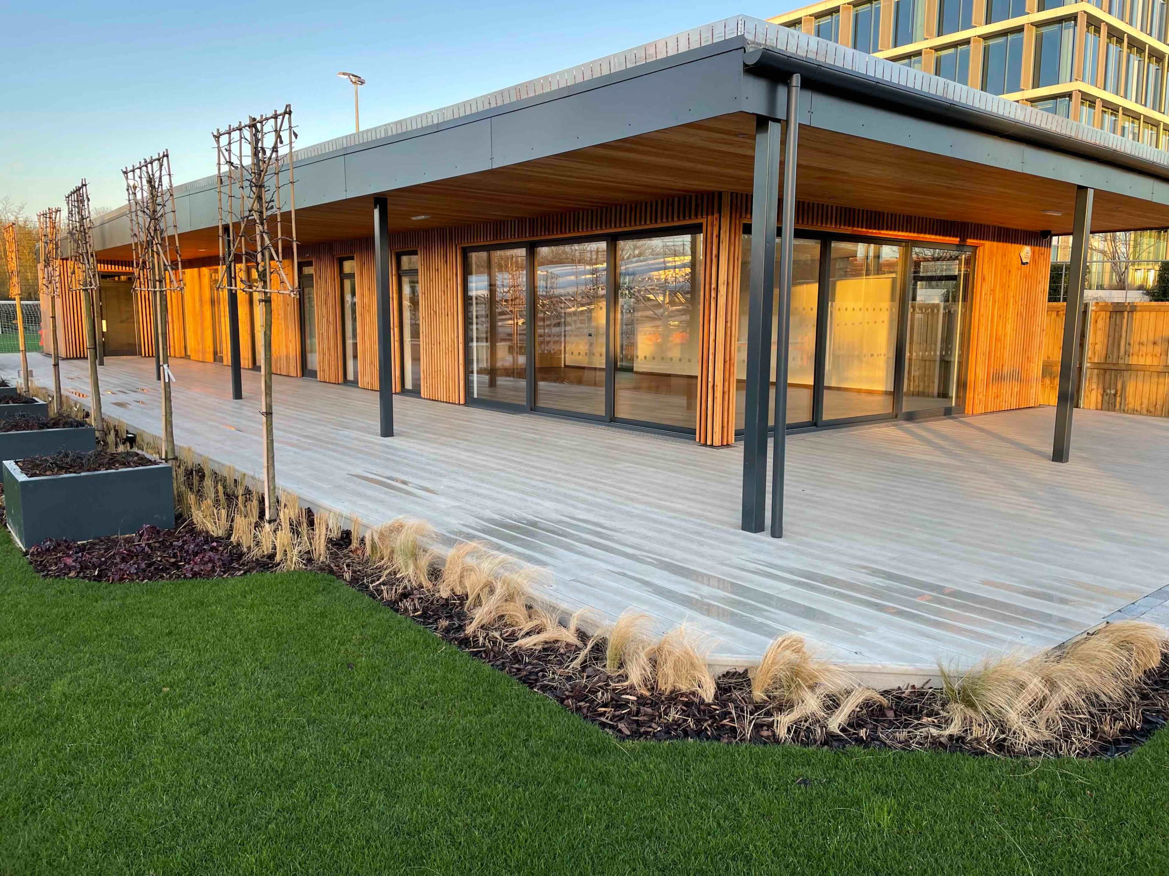 A modern building with wooden paneling and large glass windows, surrounded by a landscaped area featuring grass and decorative plants.