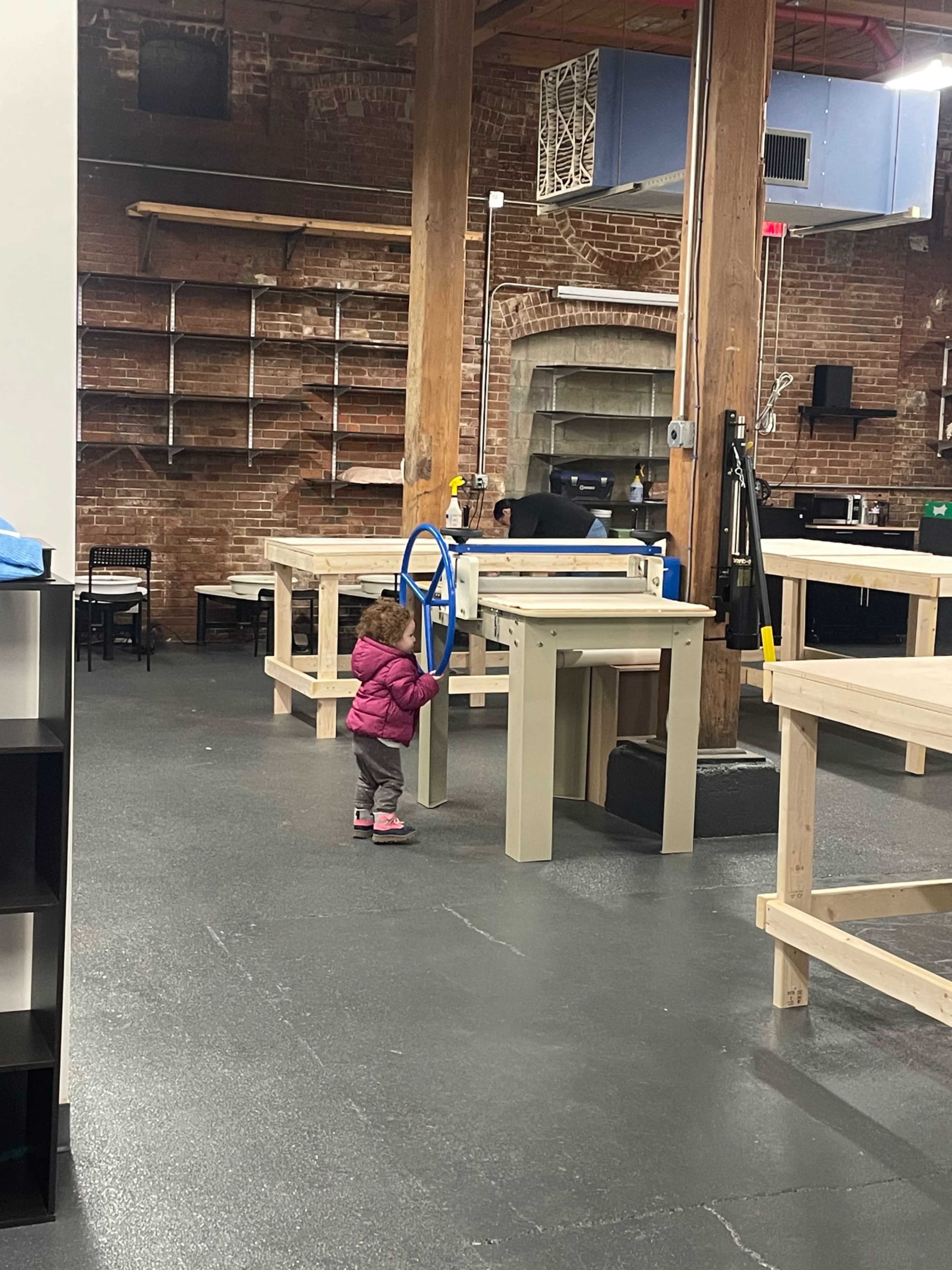 A toddler stands beside a blue wheel in a spacious workshop with wooden tables and exposed brick walls.