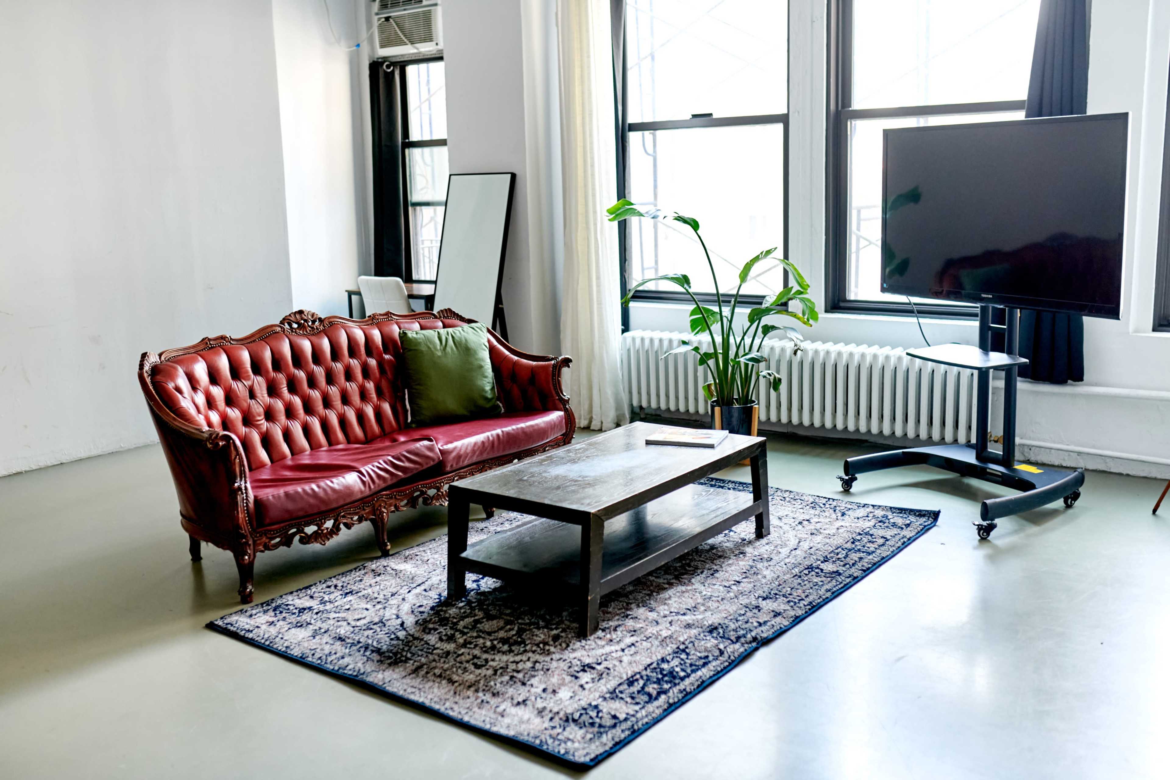 A living room setting with a red tufted sofa, a wooden coffee table on a patterned rug, and a large television on a mobile stand beside a potted plant near a window.