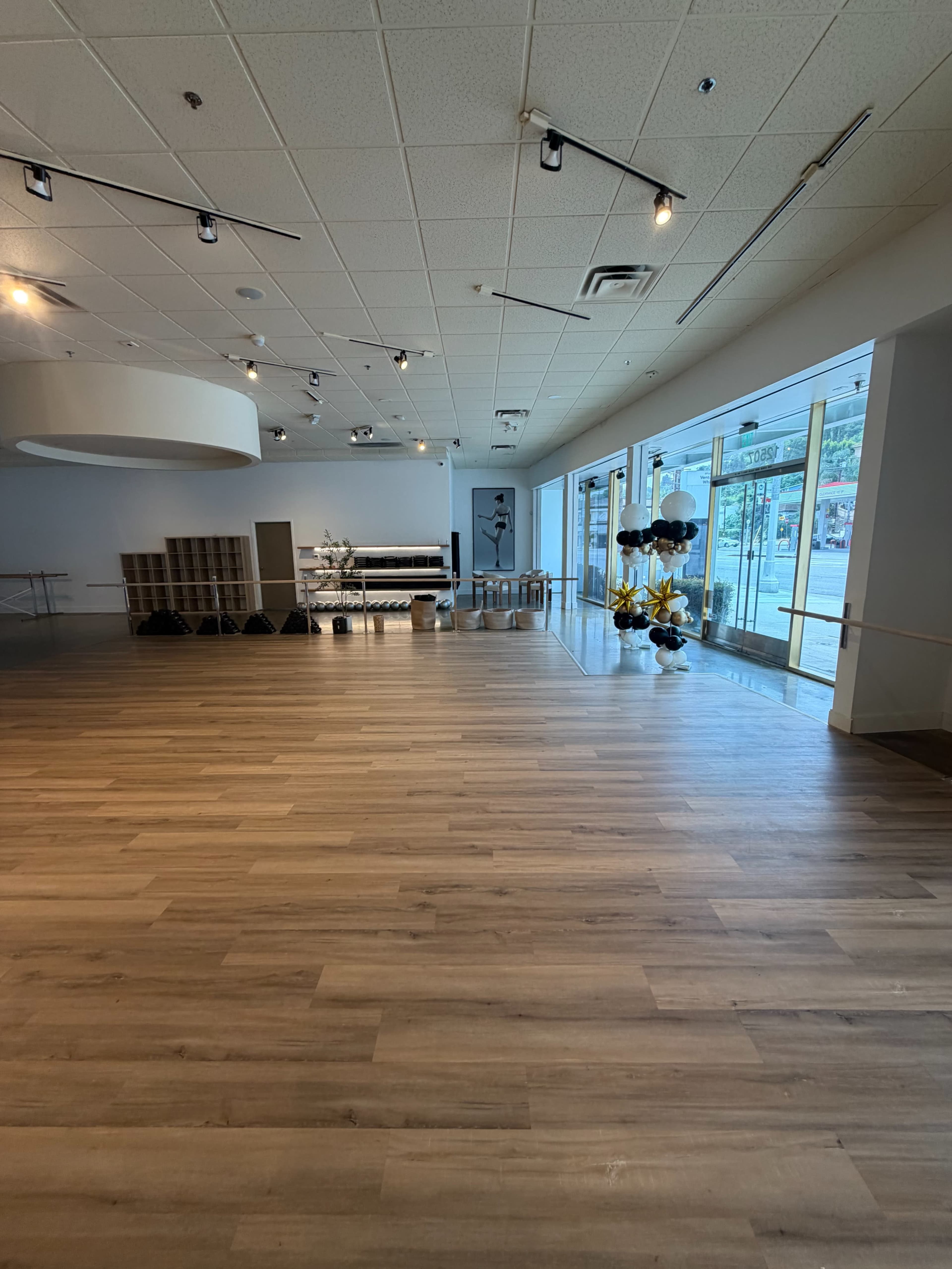The image shows a spacious, empty dance studio with wooden flooring and large windows allowing natural light to enter.
