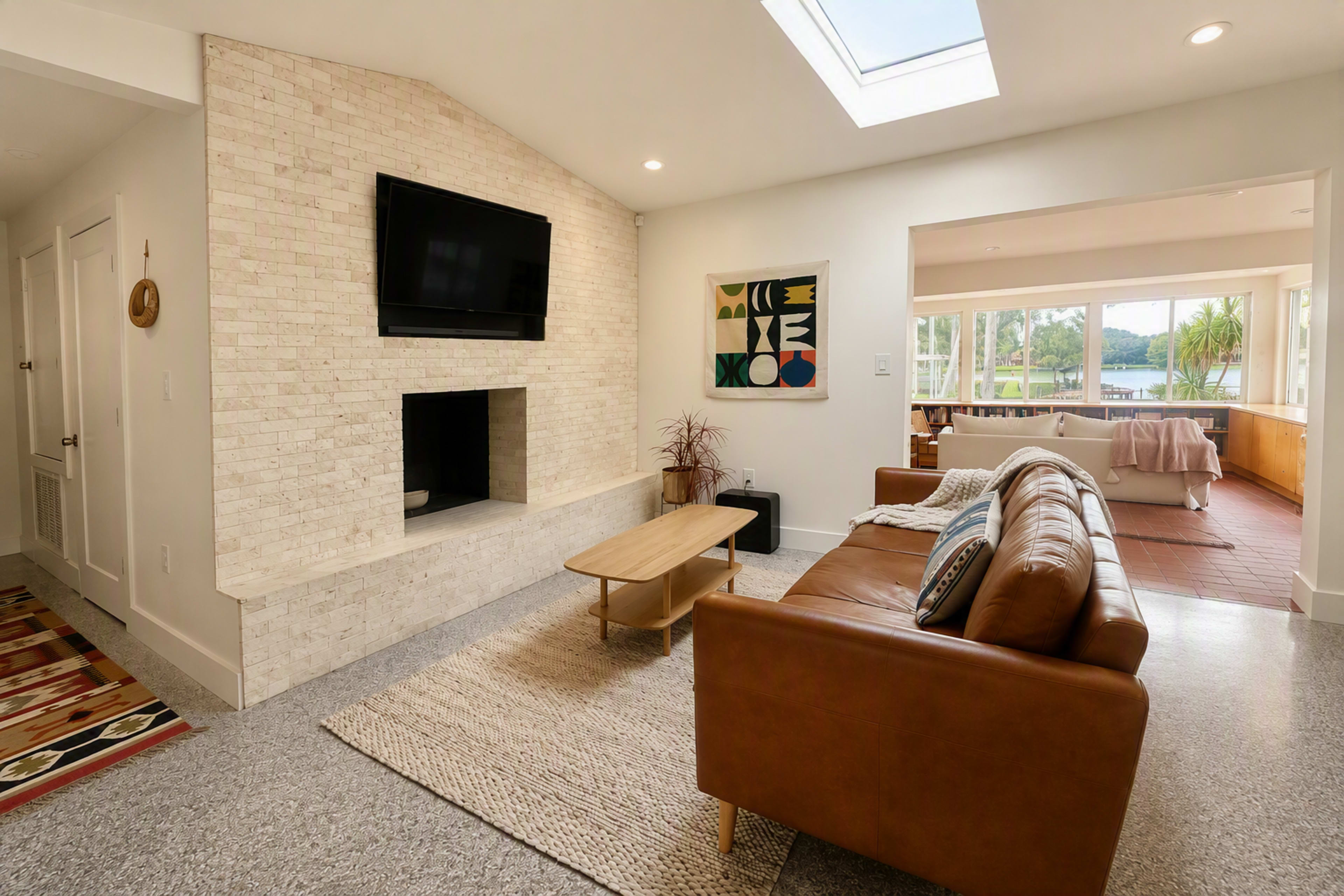 The image shows a modern living room with a beige brick wall featuring a mounted TV, a brown leather sofa, a light wooden coffee table, and large windows that let in natural light.