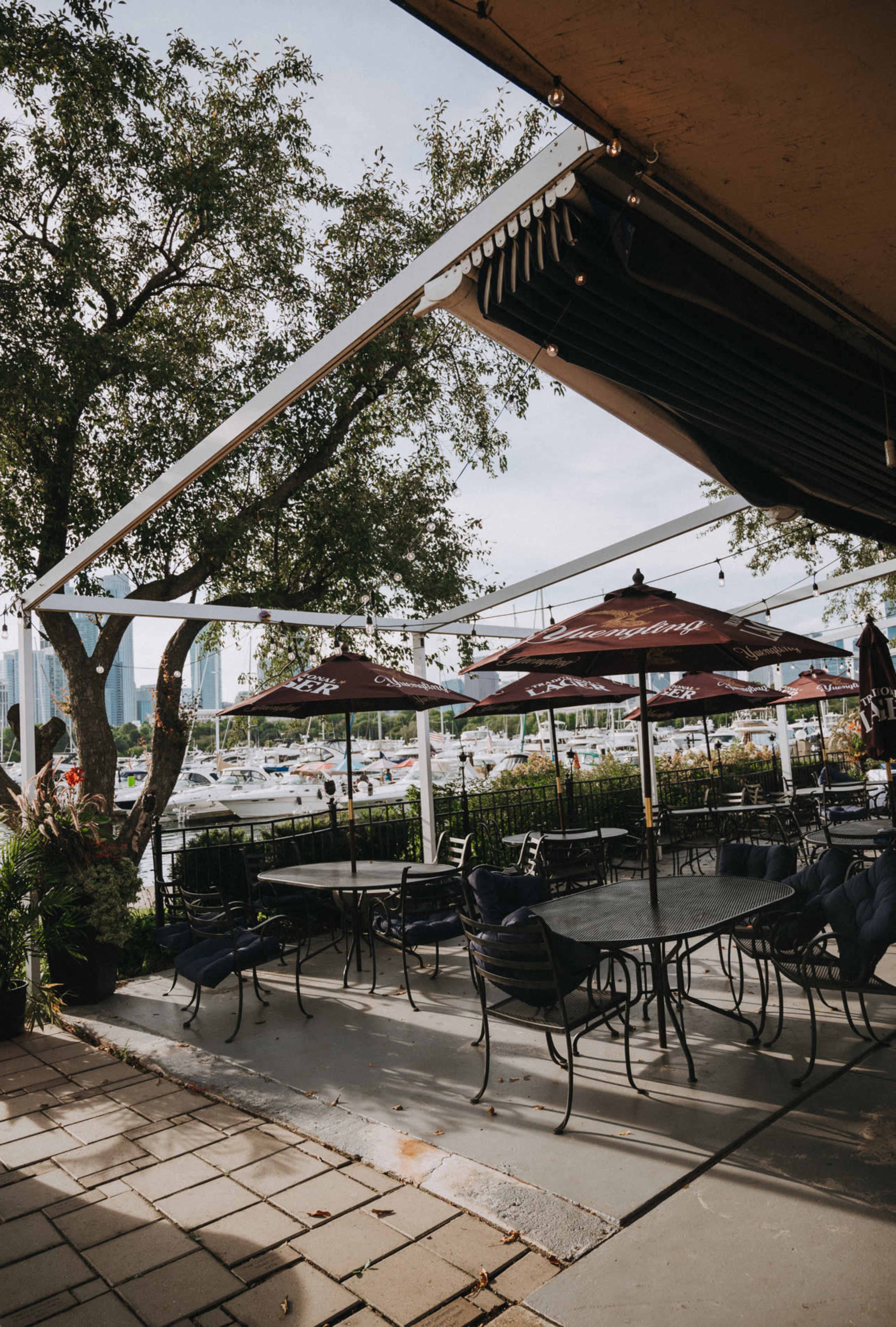 The image shows a picturesque outdoor dining area with metal tables and chairs, shaded by umbrellas, adjacent to a marina filled with boats.