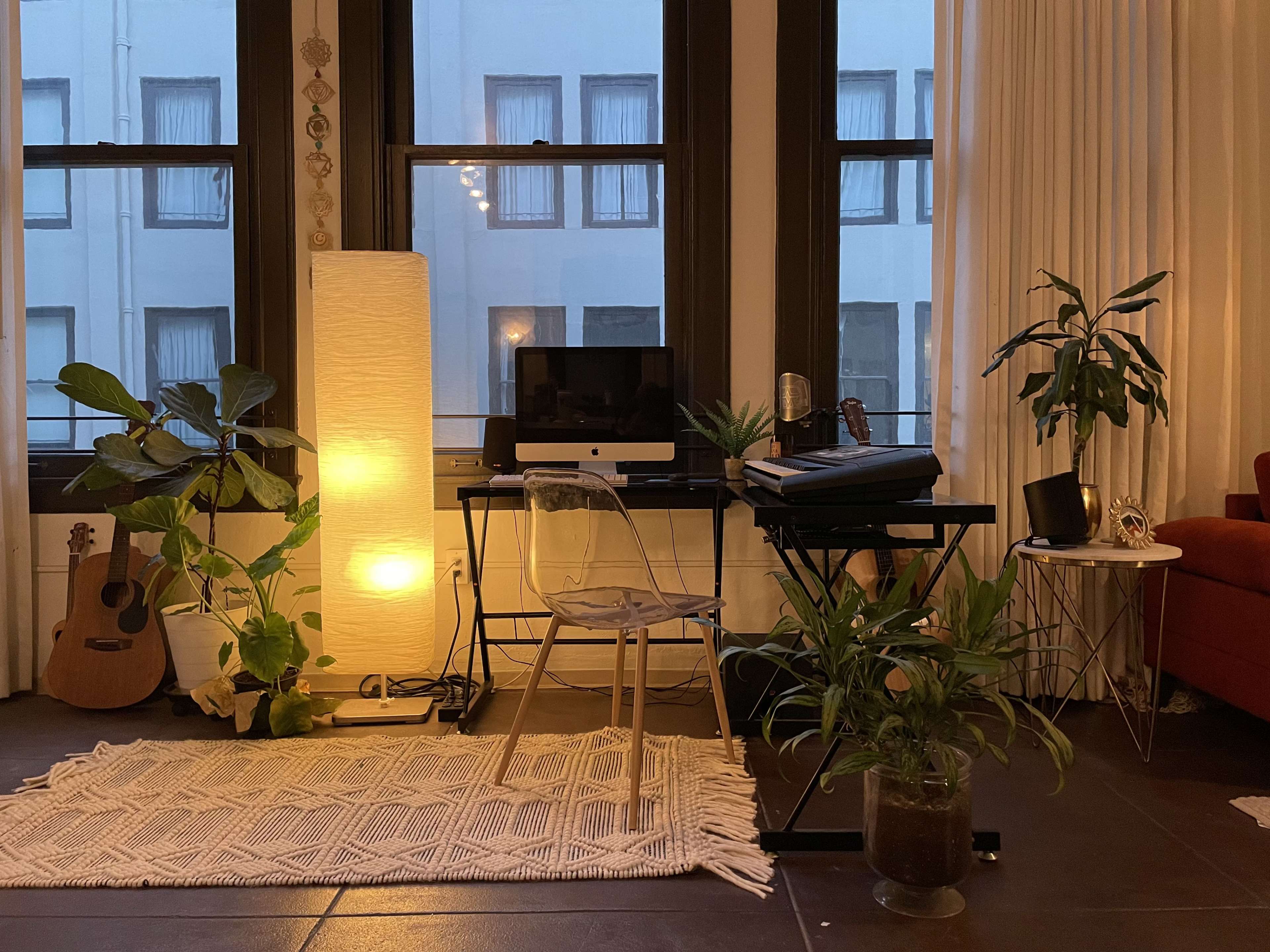 A small workspace with a computer desk, a chair, plants, a lamp, and a guitar, situated near a window.