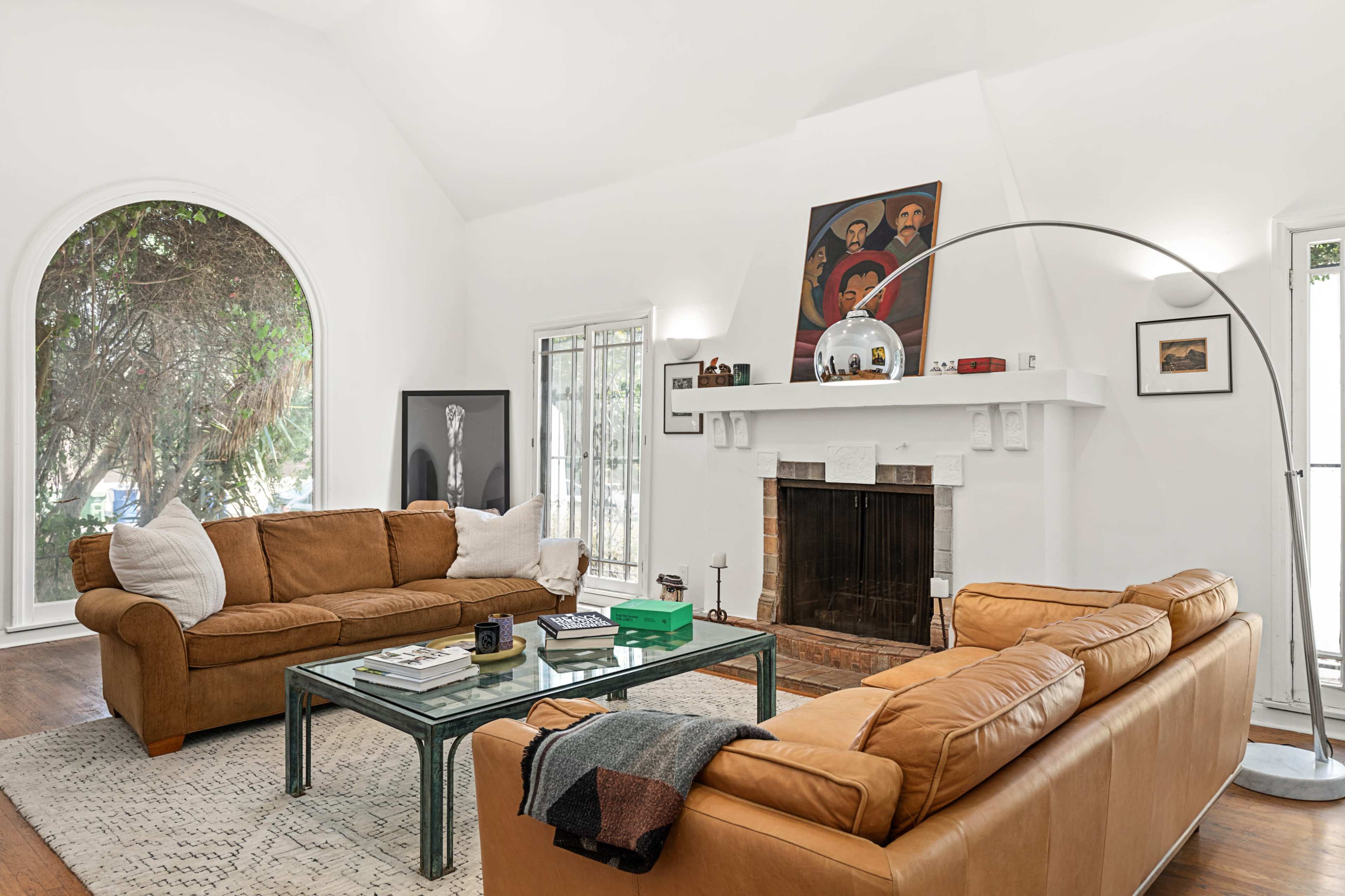 A bright living room with two brown leather sofas facing a glass coffee table and a fireplace, accented by a large painting above the mantel.