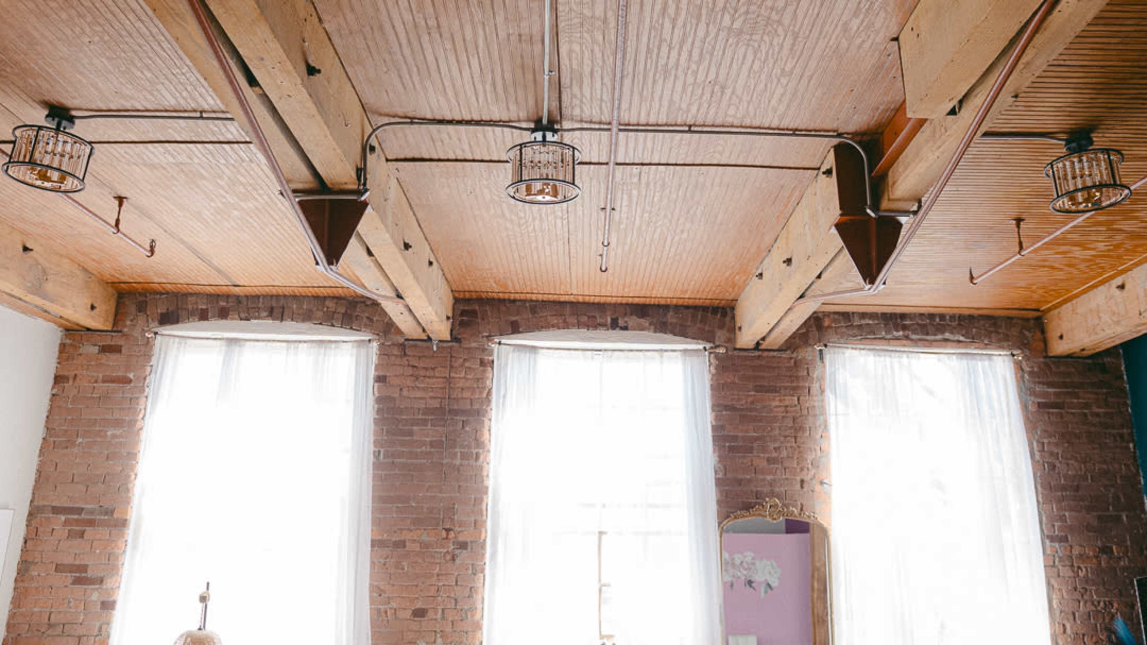 A room with exposed wooden beams, metal light fixtures, and large windows draped with white curtains against a brick wall.
