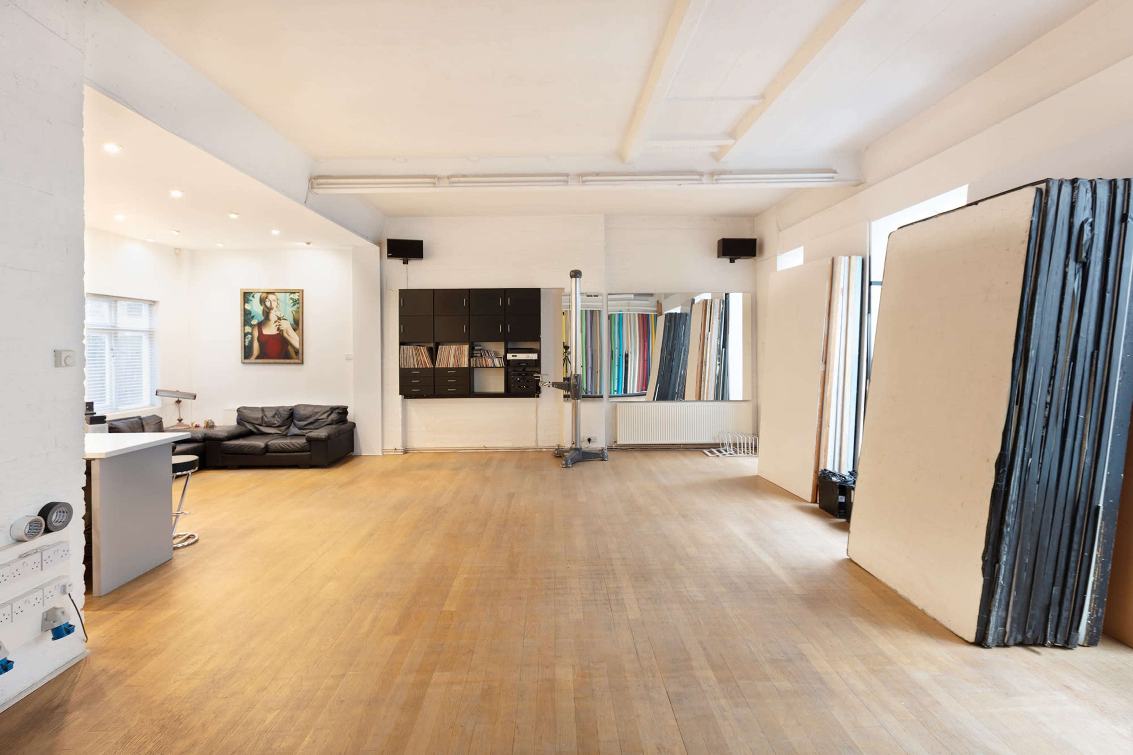 A spacious, well-lit studio with wooden flooring, featuring black furniture, large mirrors, and an array of colored panels against the wall.