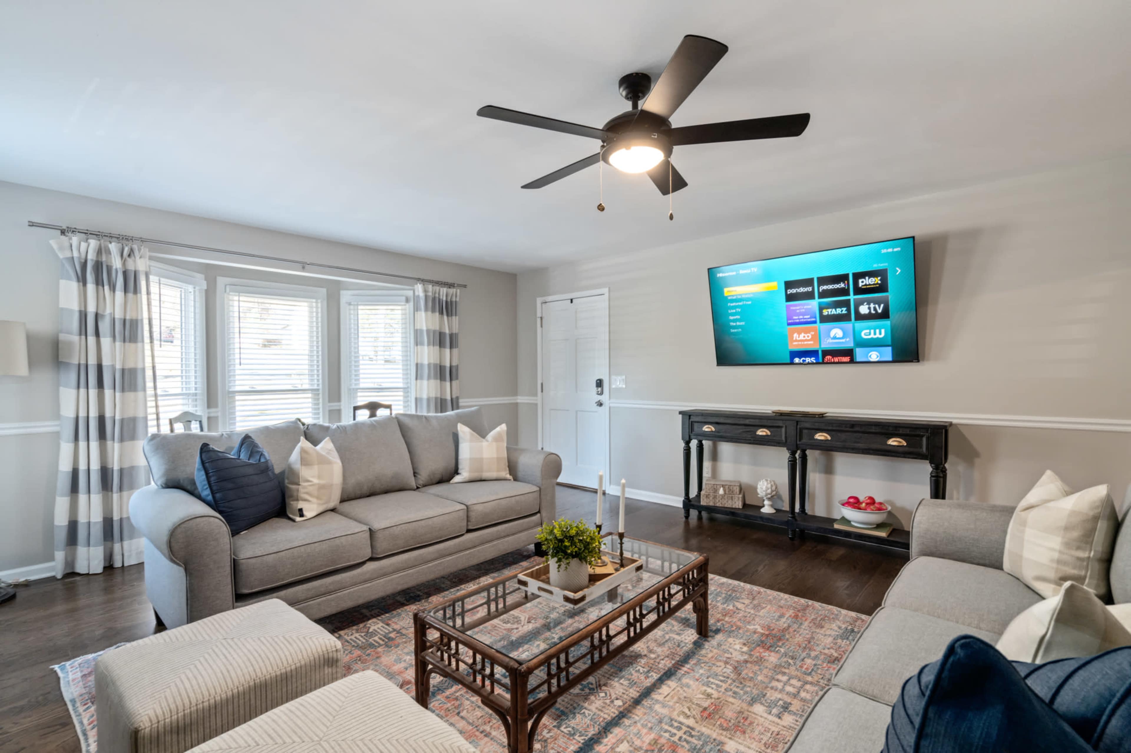 A well-furnished living room features a gray sofa set, a coffee table, a television mounted on the wall, and large windows dressed with curtains.