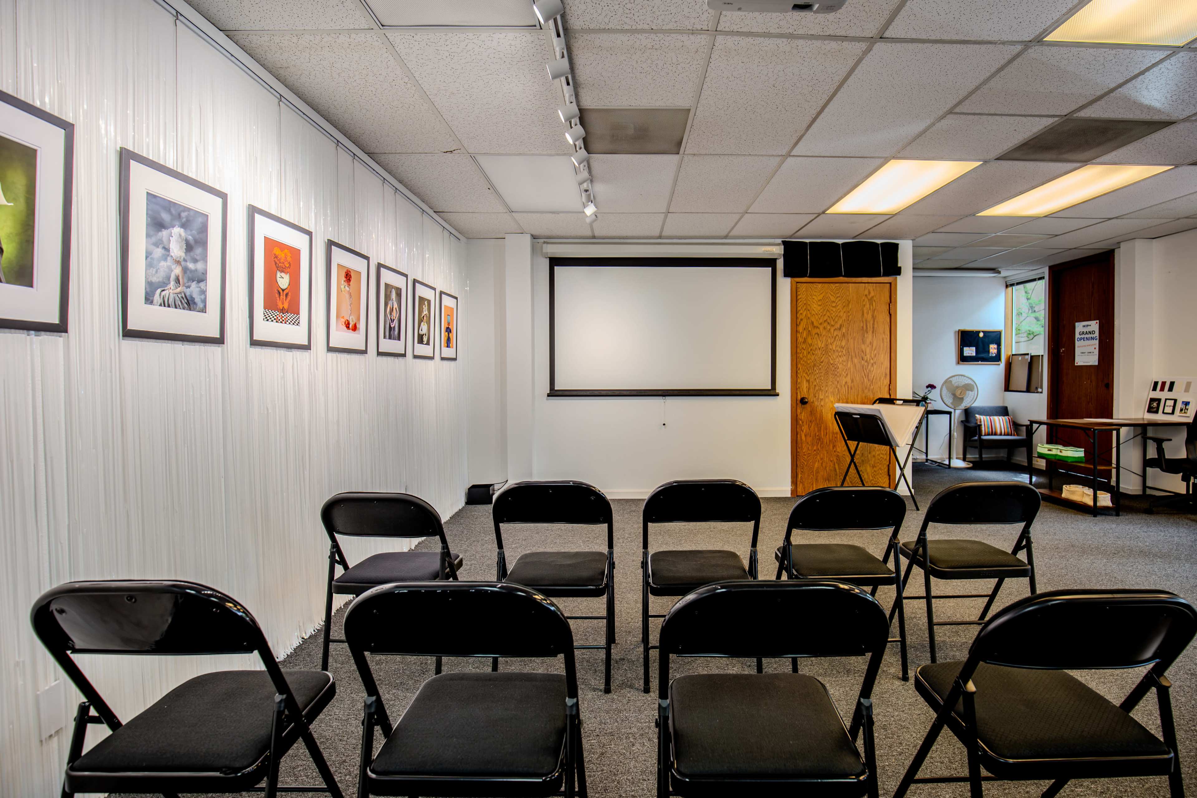 The image shows a seminar room with black folding chairs arranged facing a blank screen, surrounded by framed artwork on the walls.