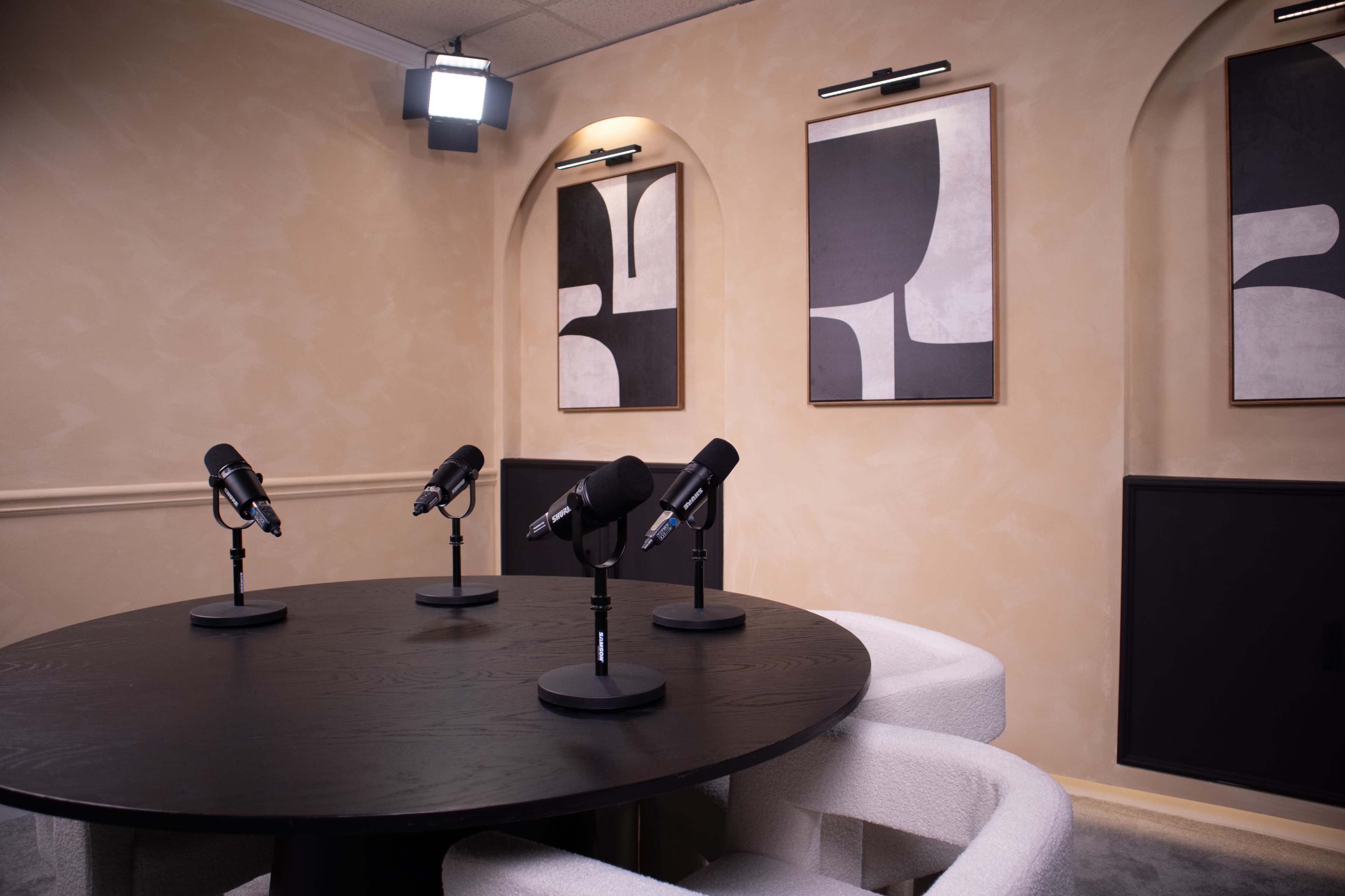 A modern podcast studio features a round black table surrounded by white chairs, with four microphones positioned at equal intervals, and abstract artwork on the walls.