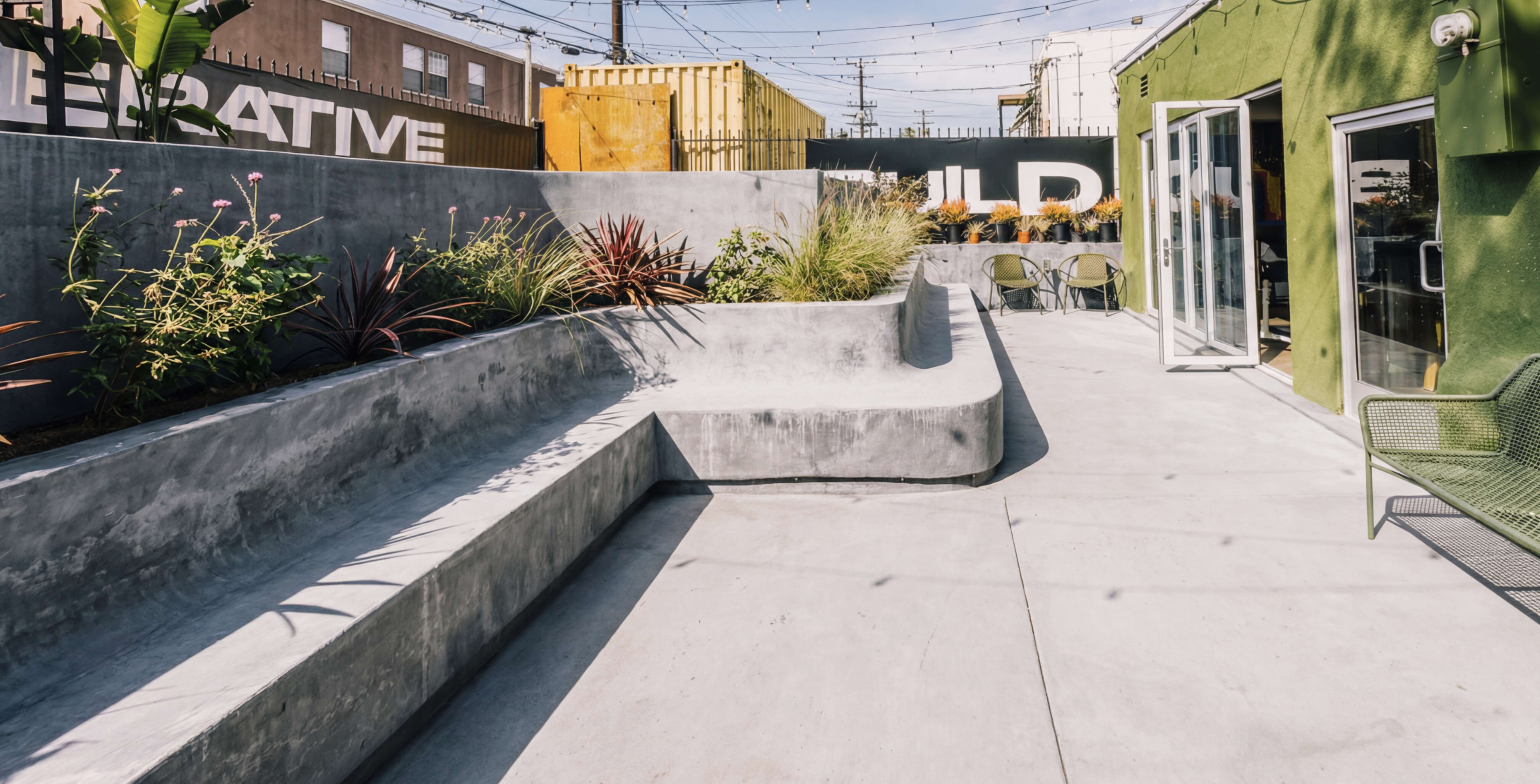 The image shows a modern outdoor space with curvilinear concrete seating areas, planters, and a green building facade in the background.