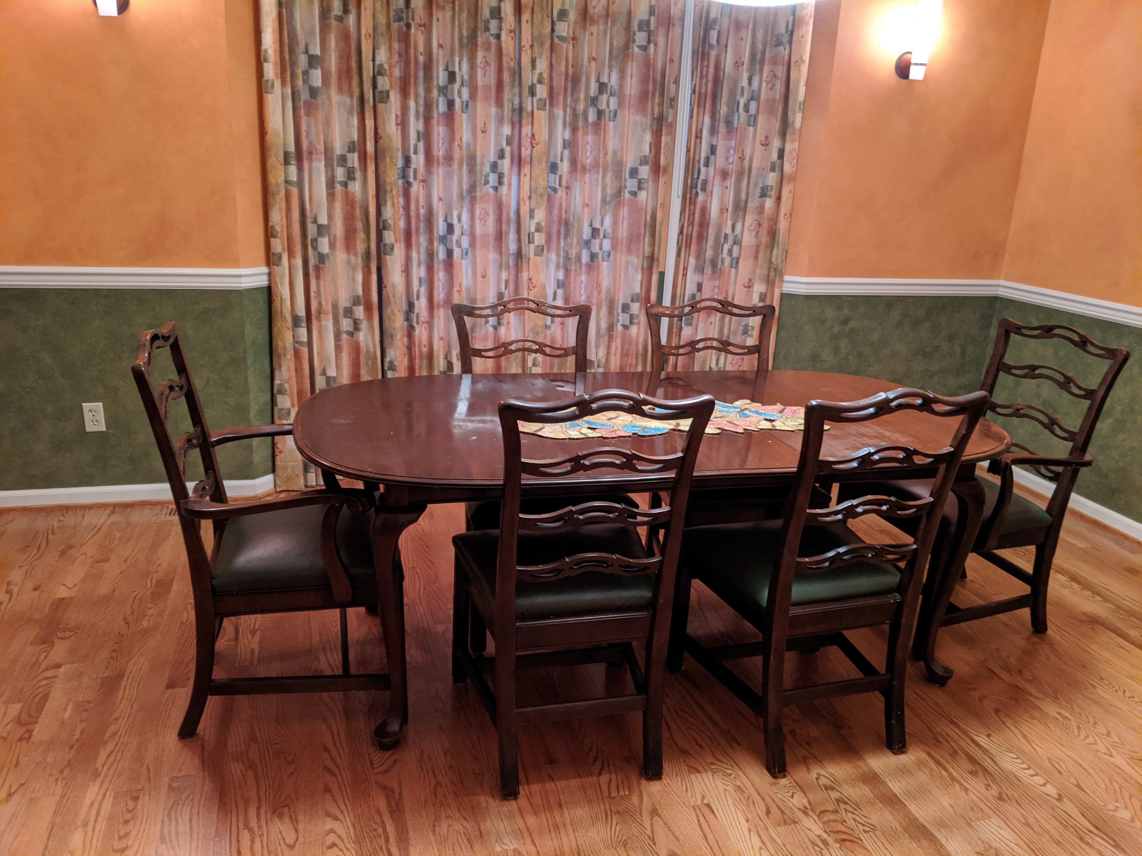 A wooden dining table surrounded by six chairs sits in a room with orange walls and patterned curtains.