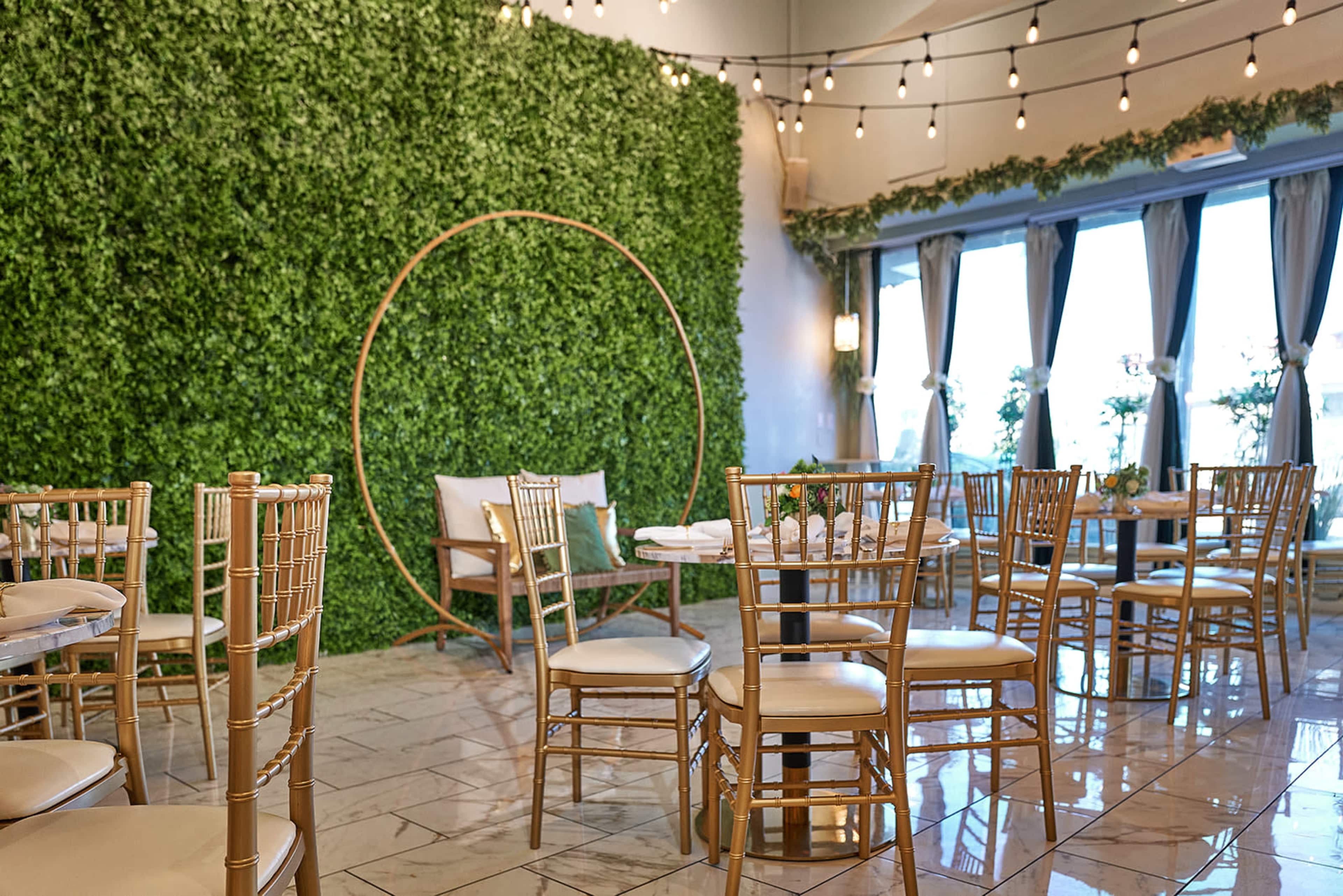 A dining area features round tables with gold chairs, a circular floral backdrop, and string lights above.
