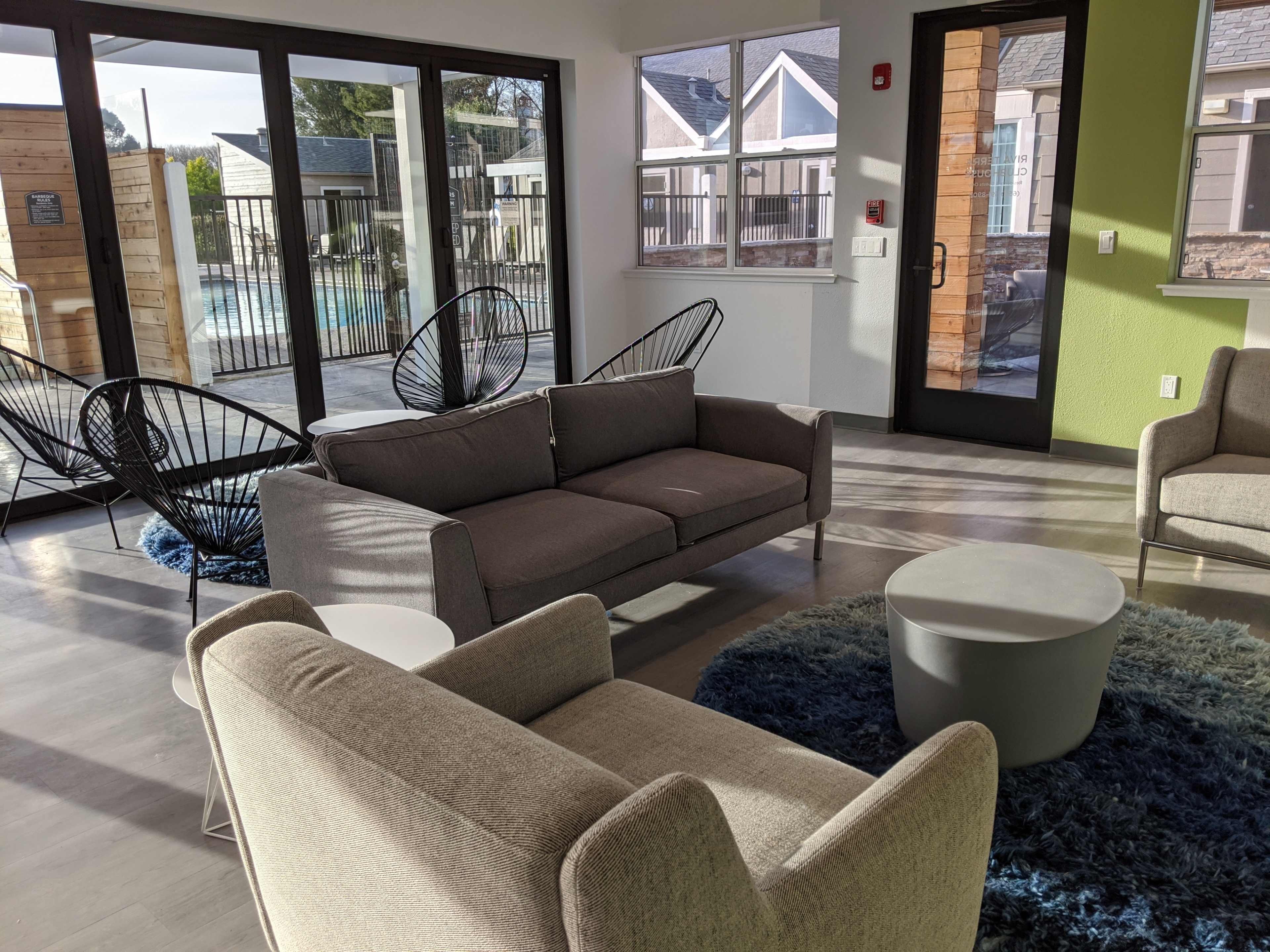 The image shows a modern lounge area with a light gray sofa, two chairs, a round coffee table, and large windows overlooking a pool.