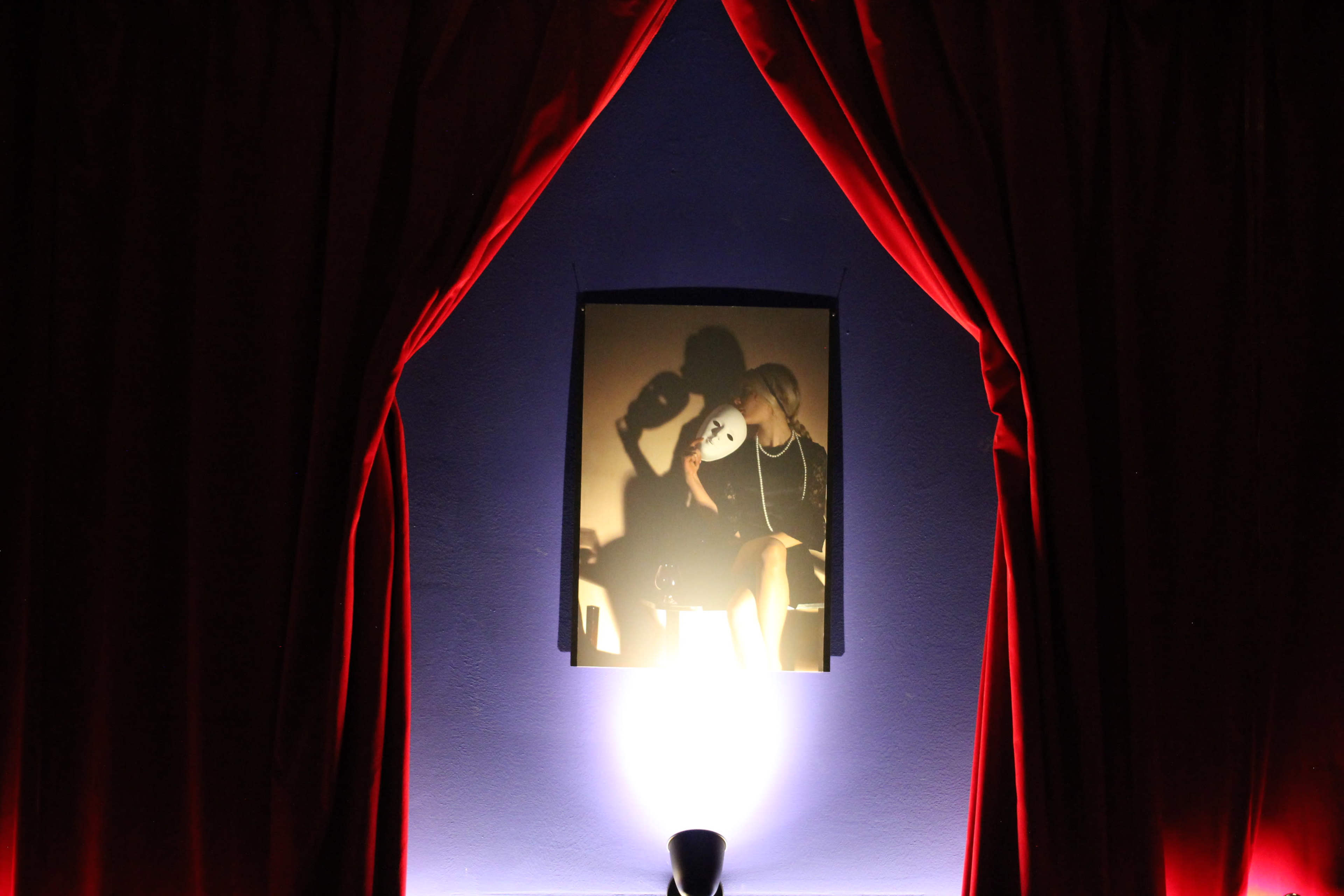 A framed picture of a person holding a mask is illuminated by a light and surrounded by red curtains.