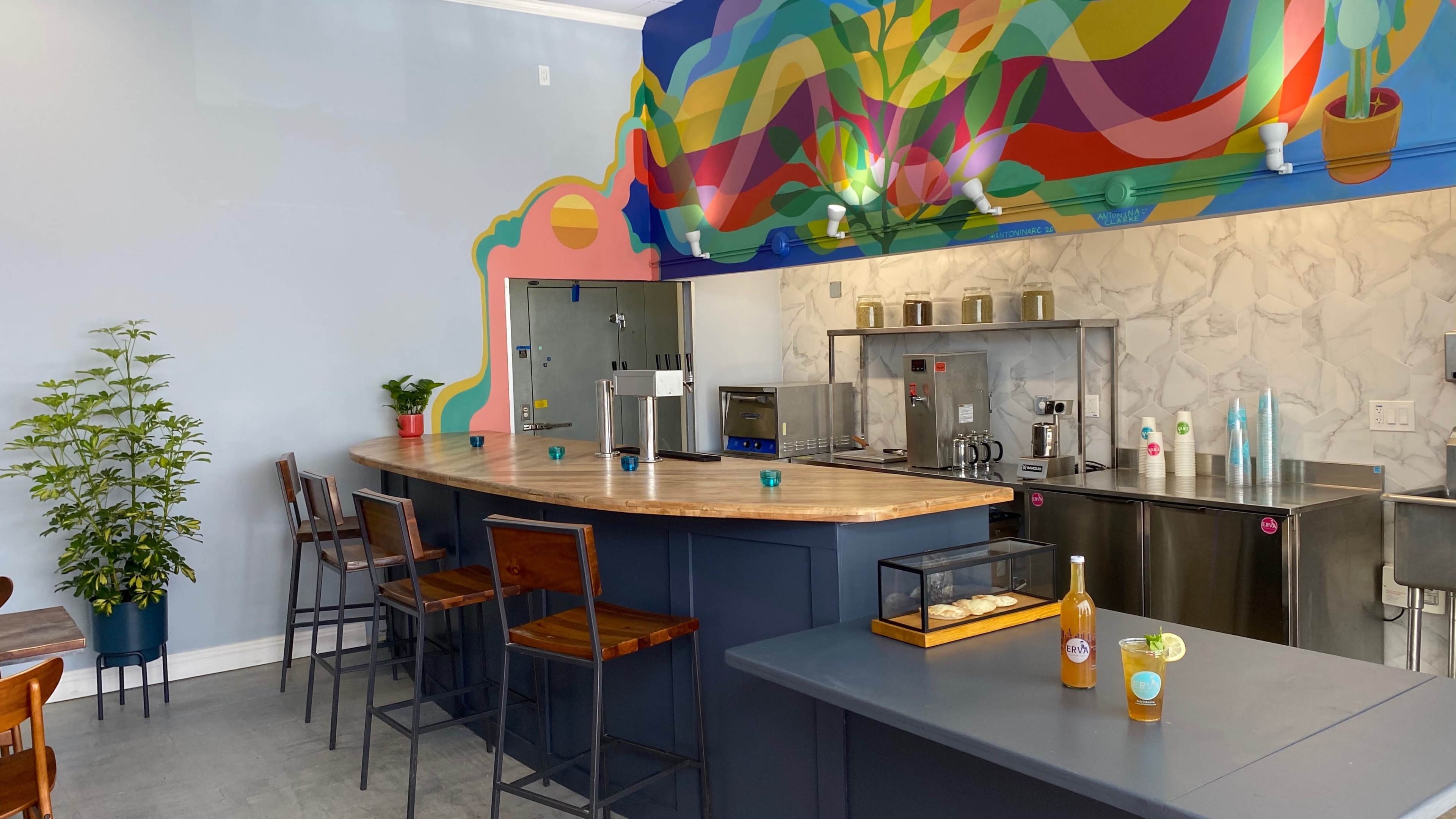 A modern cafe interior featuring a curved wooden bar with high stools, colorful mural artwork on the wall, and various kitchen appliances in a stainless steel setup.
