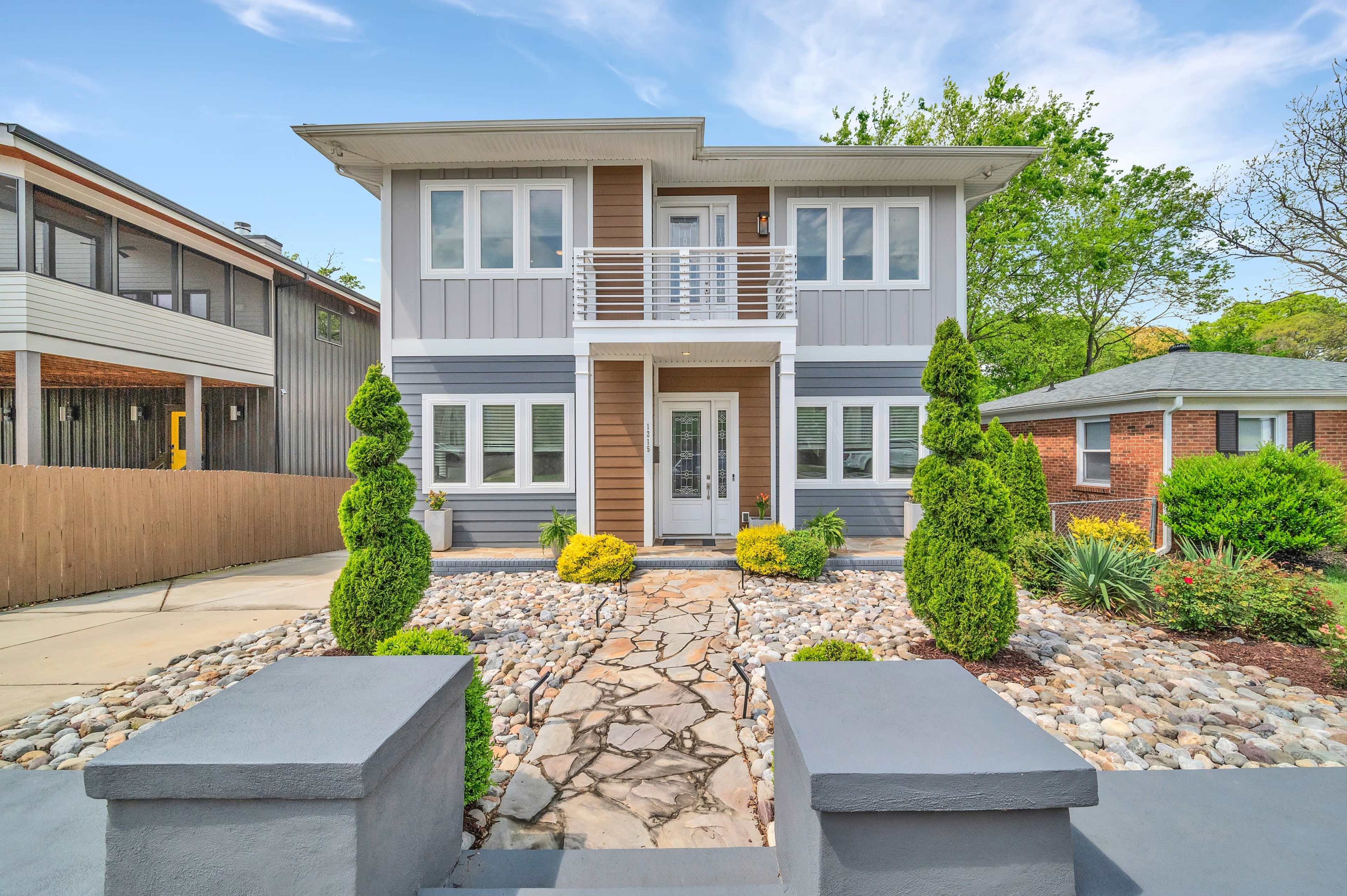 A two-story house with a gray and white exterior features a stone path leading to the front door, flanked by neatly trimmed bushes and landscaping.