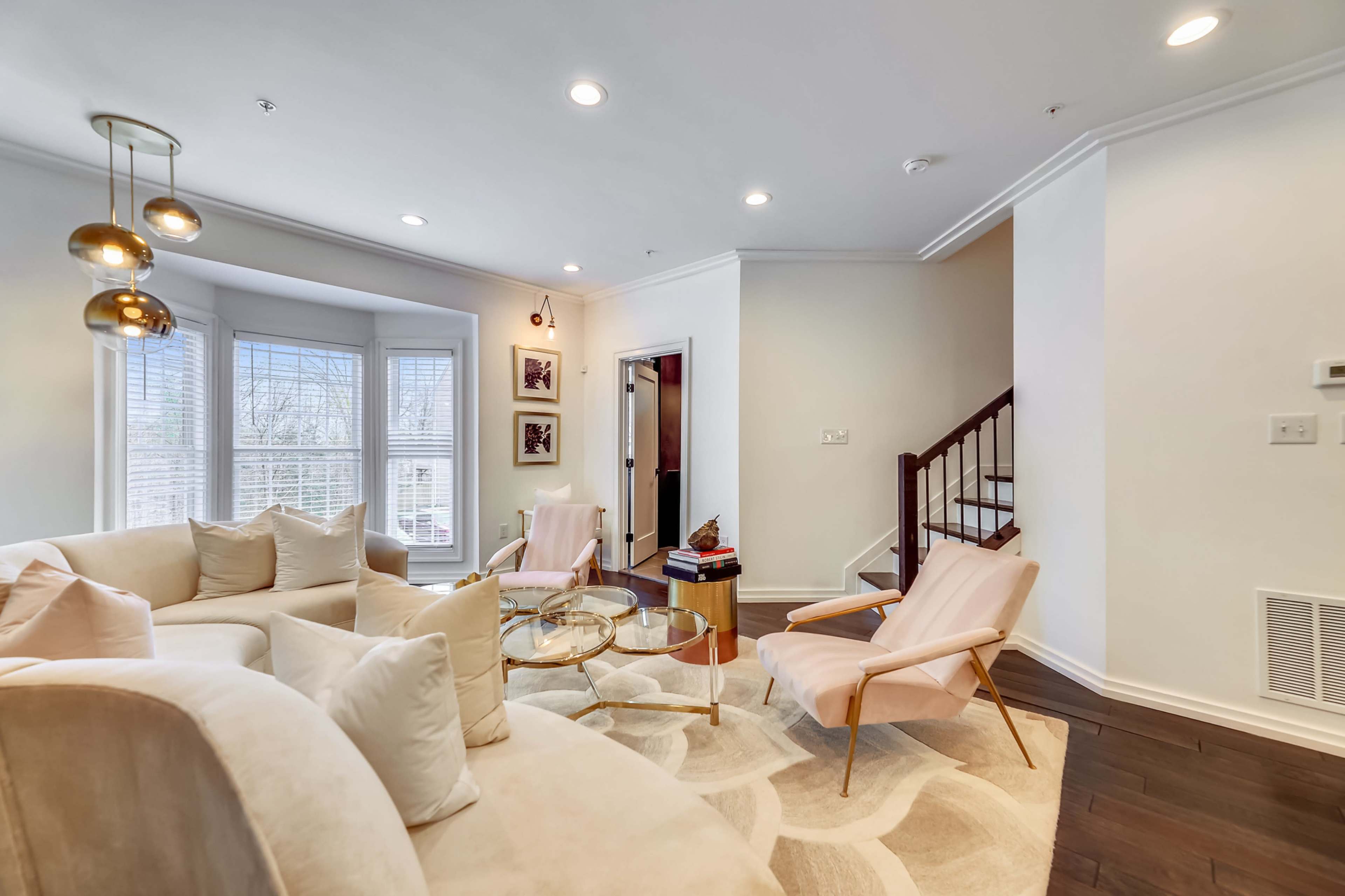 A modern living room with a curved beige sectional sofa, two light pink armchairs, a round glass coffee table, and a staircase in the background.