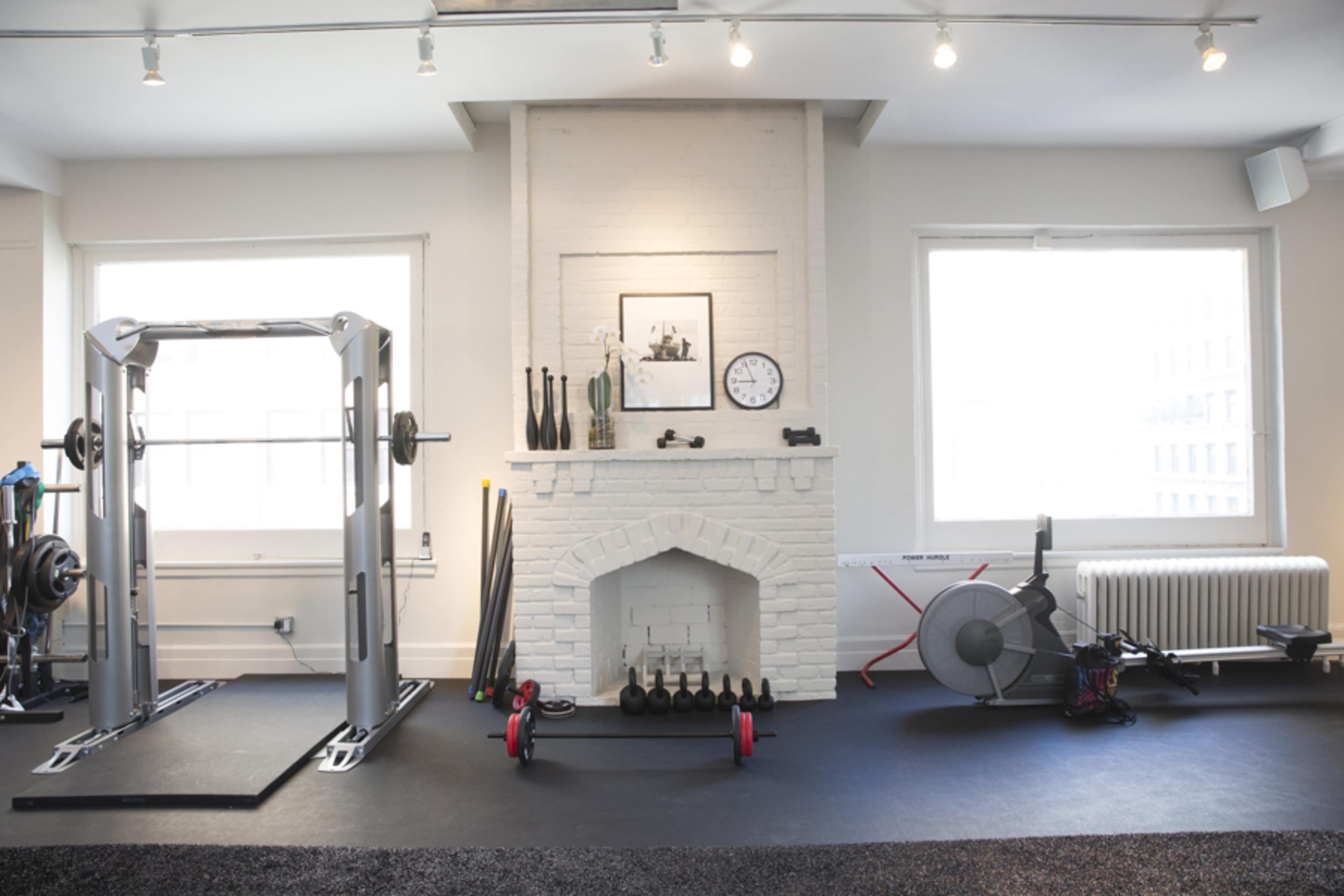 The image shows a bright gym with a white brick fireplace, weightlifting equipment, and exercise accessories arranged along the walls.