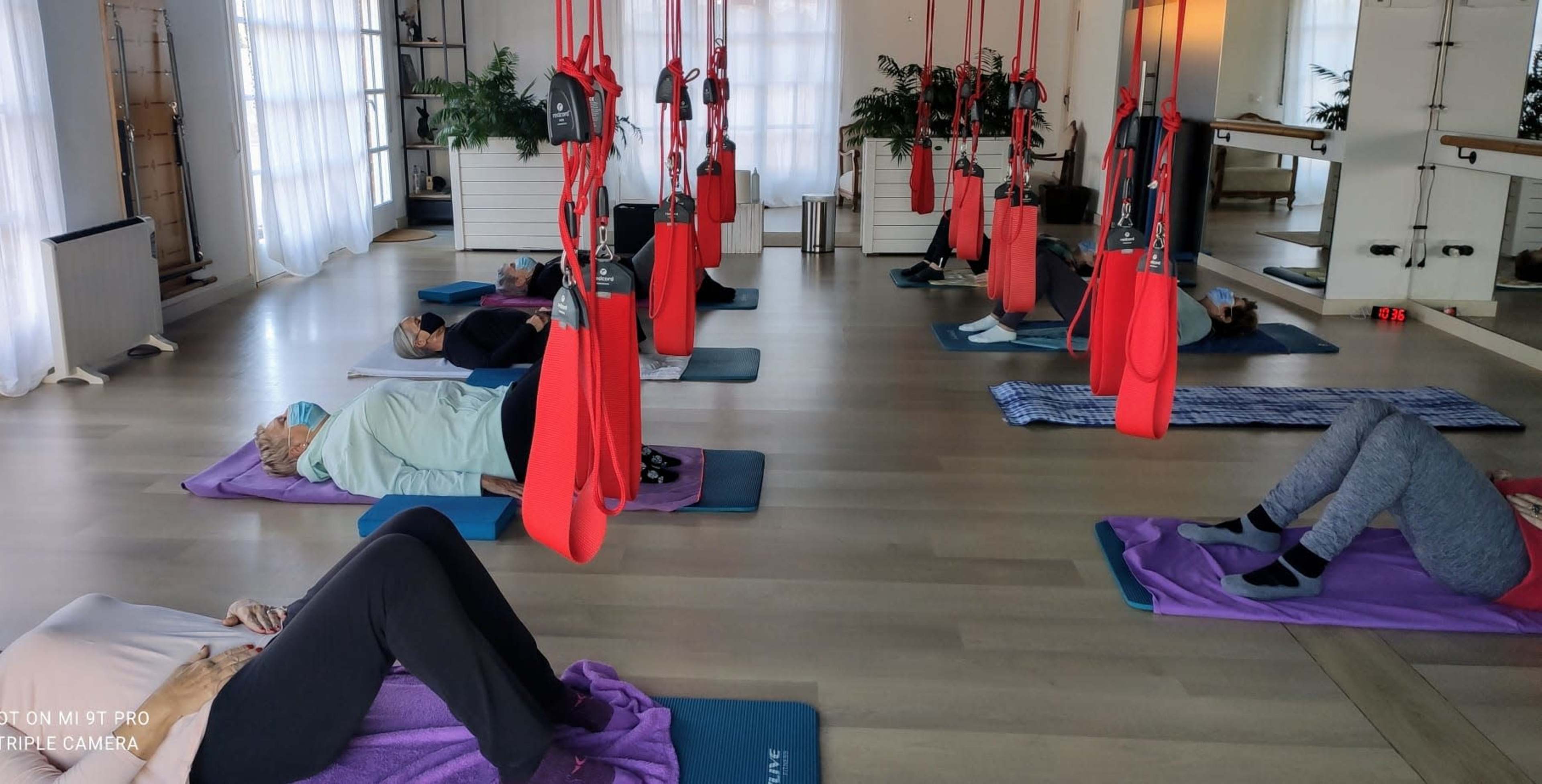A group of people lie on exercise mats in a studio equipped with red suspension straps and mirrors.