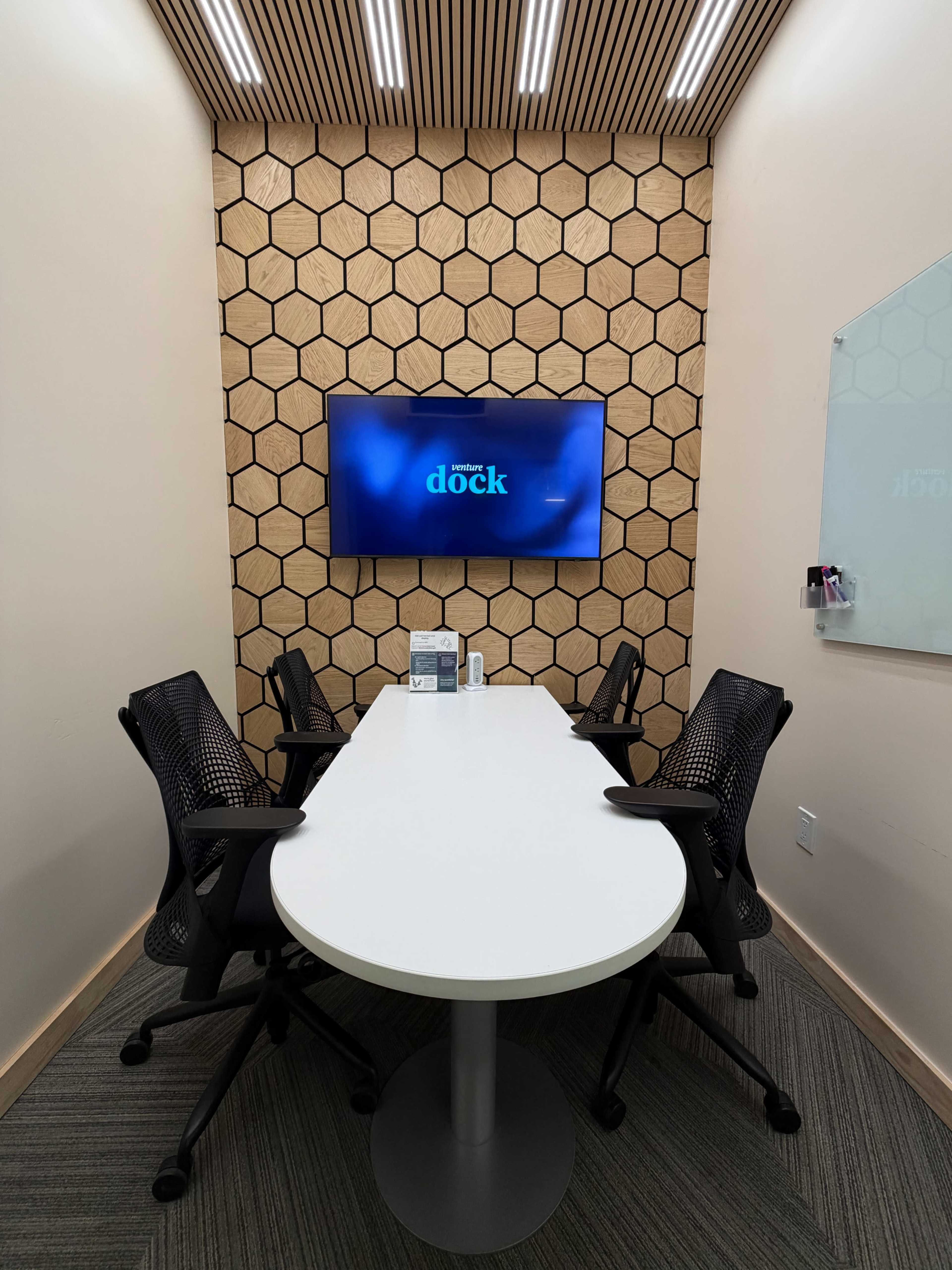 The image shows a small meeting room with a hexagonal wood accent wall, a large screen displaying "dock," and four ergonomic chairs around a white oval table.