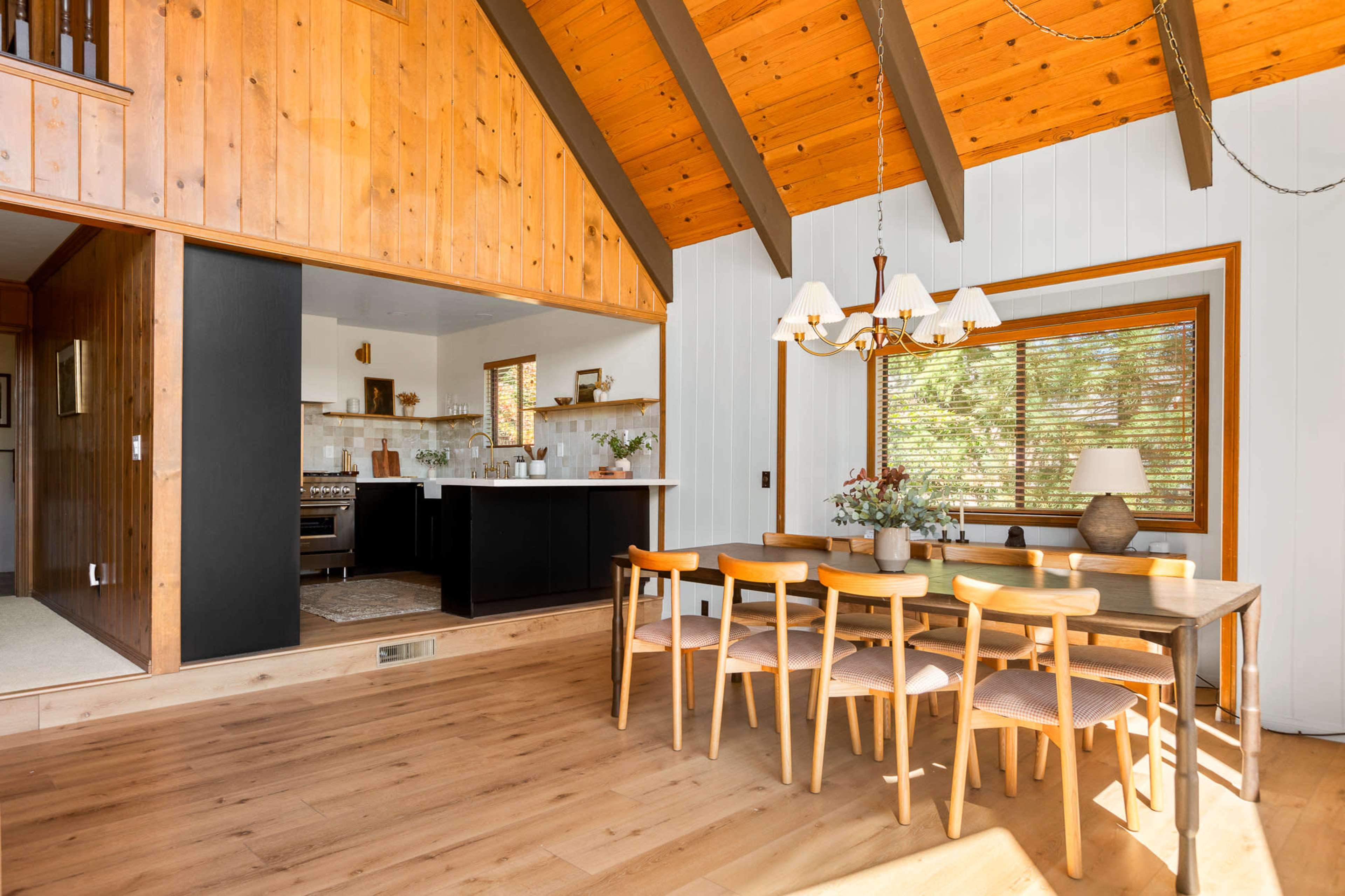 A dining area features a wooden table with chairs, illuminated by a chandelier, and opens to a kitchen in a cabin-style room with wooden accents.