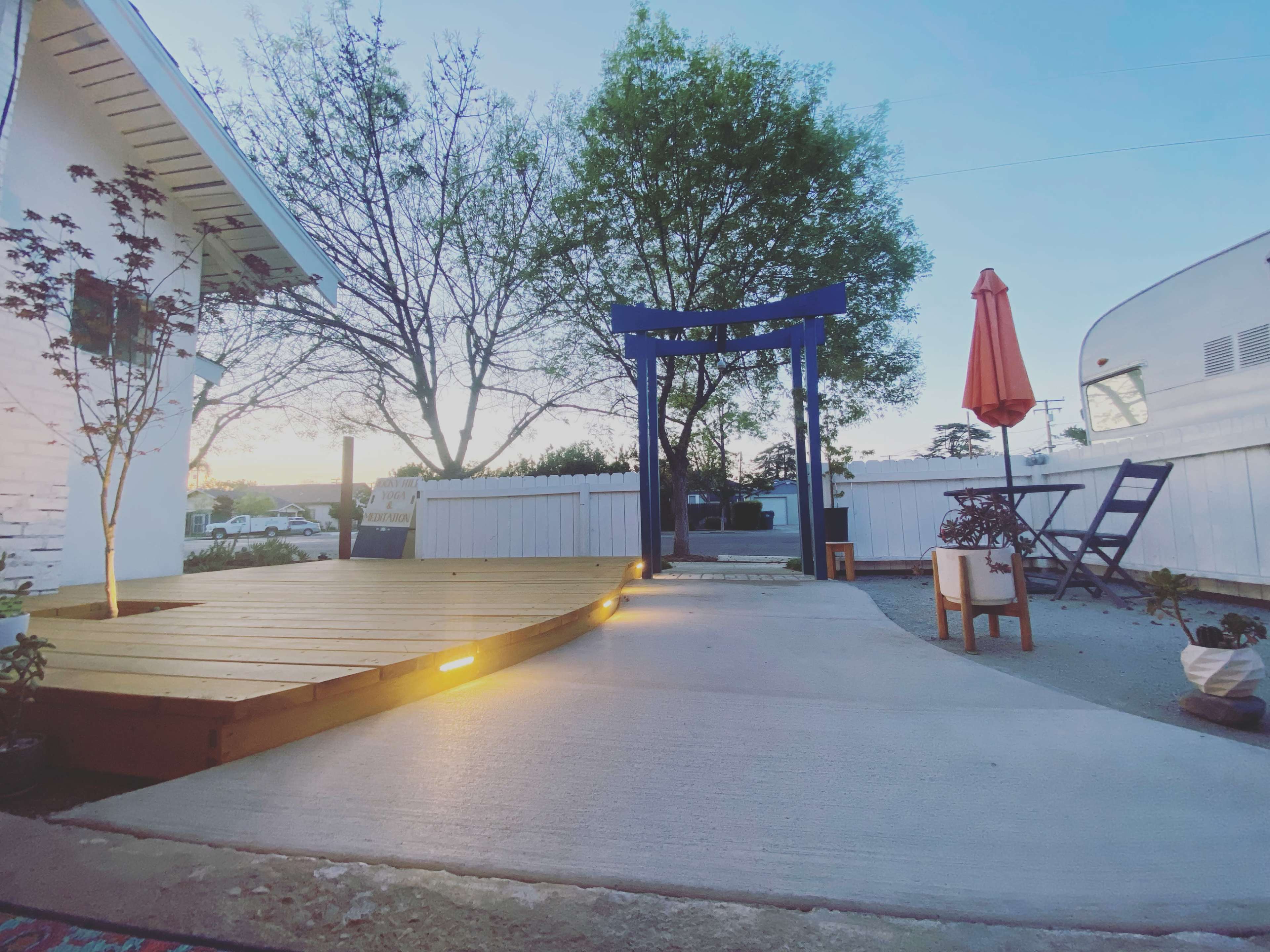 A backyard area featuring a wooden deck, a blue torii gate, an umbrella-shaded table, and a concrete pathway leading to the entrance.