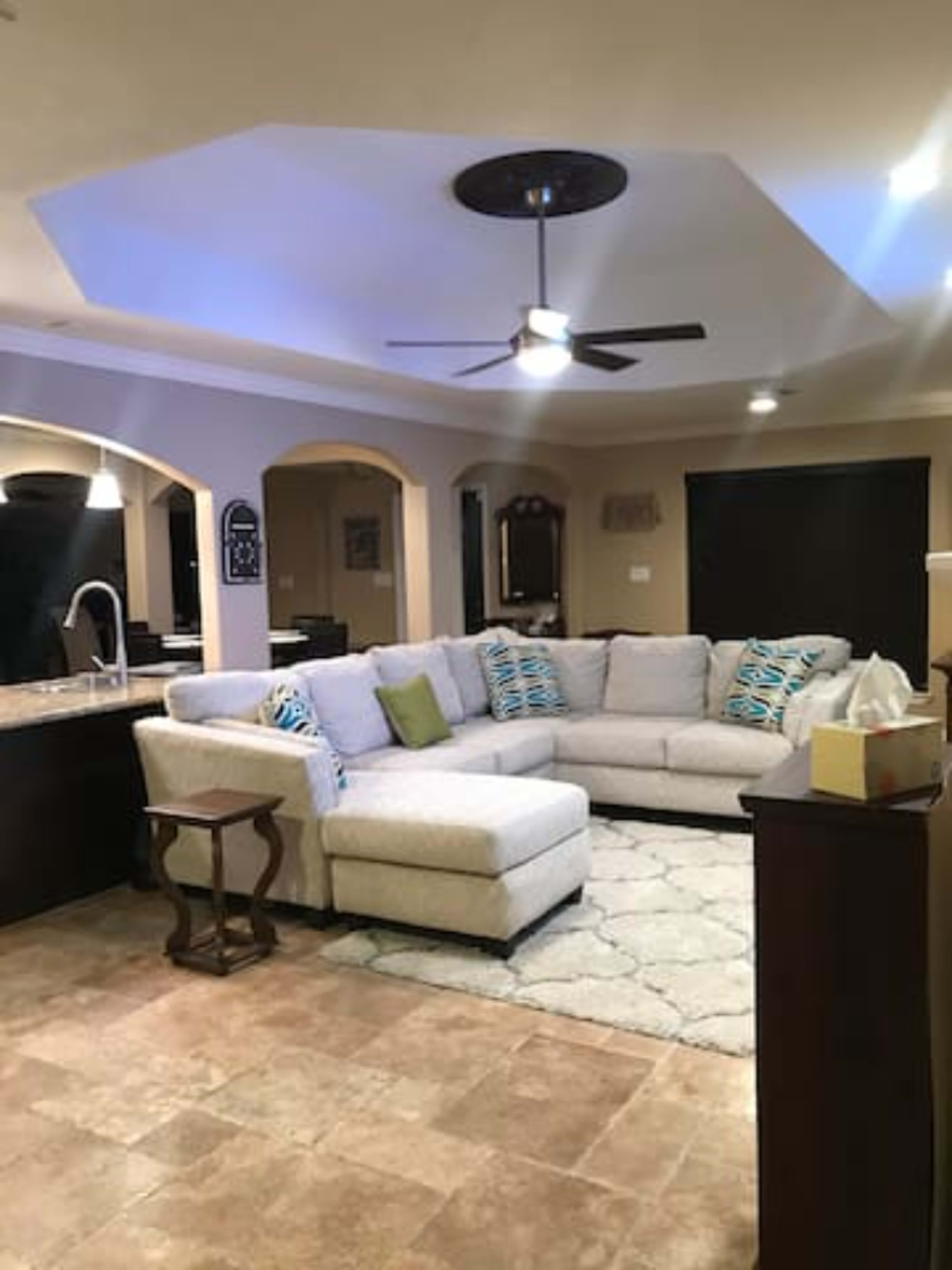 A living room with a large sectional sofa, a stone-tiled floor, and a lighted ceiling fan.