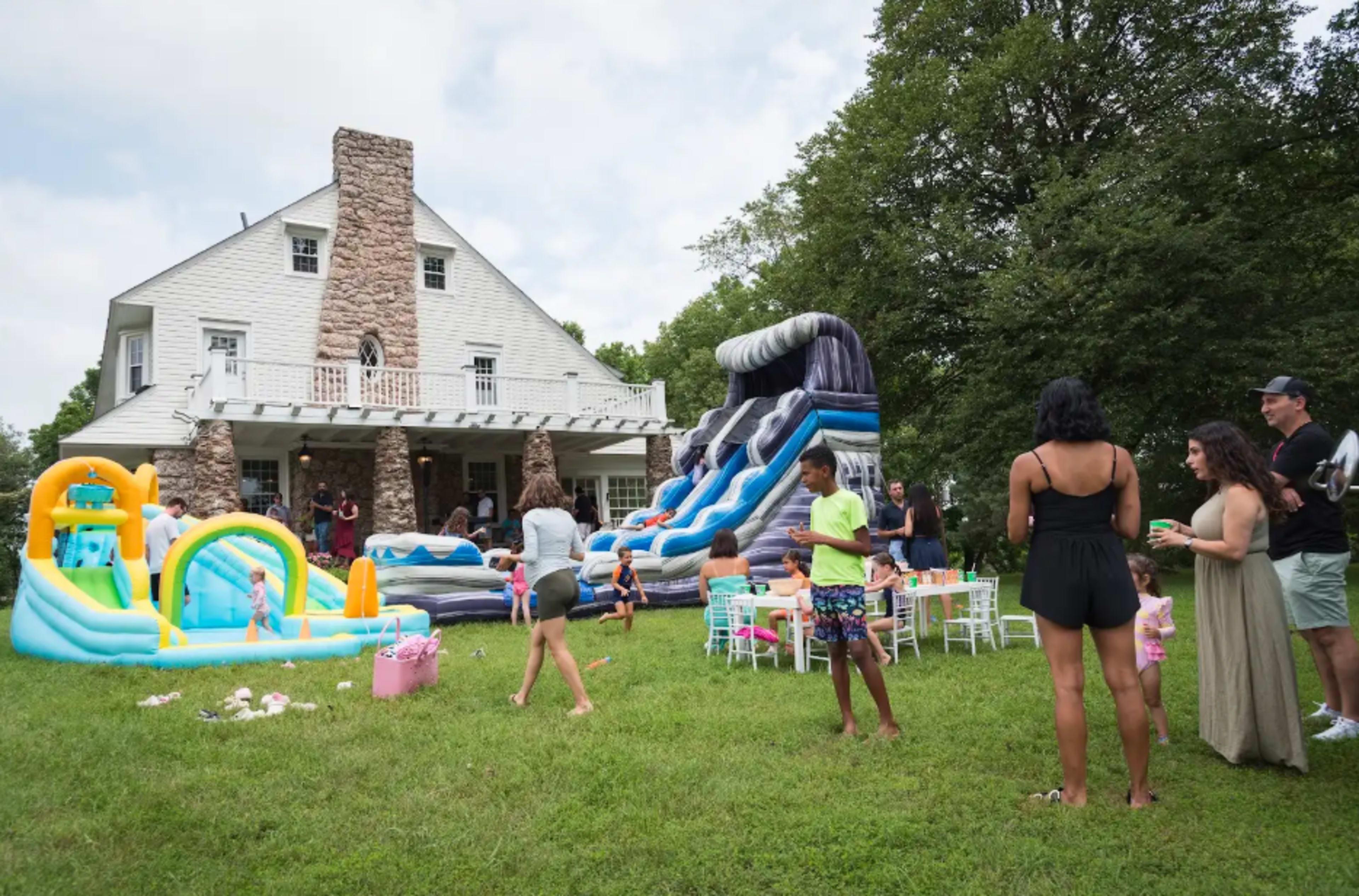A backyard party features inflatable water slides and pools, with guests socializing and children playing on the grass near a large house.