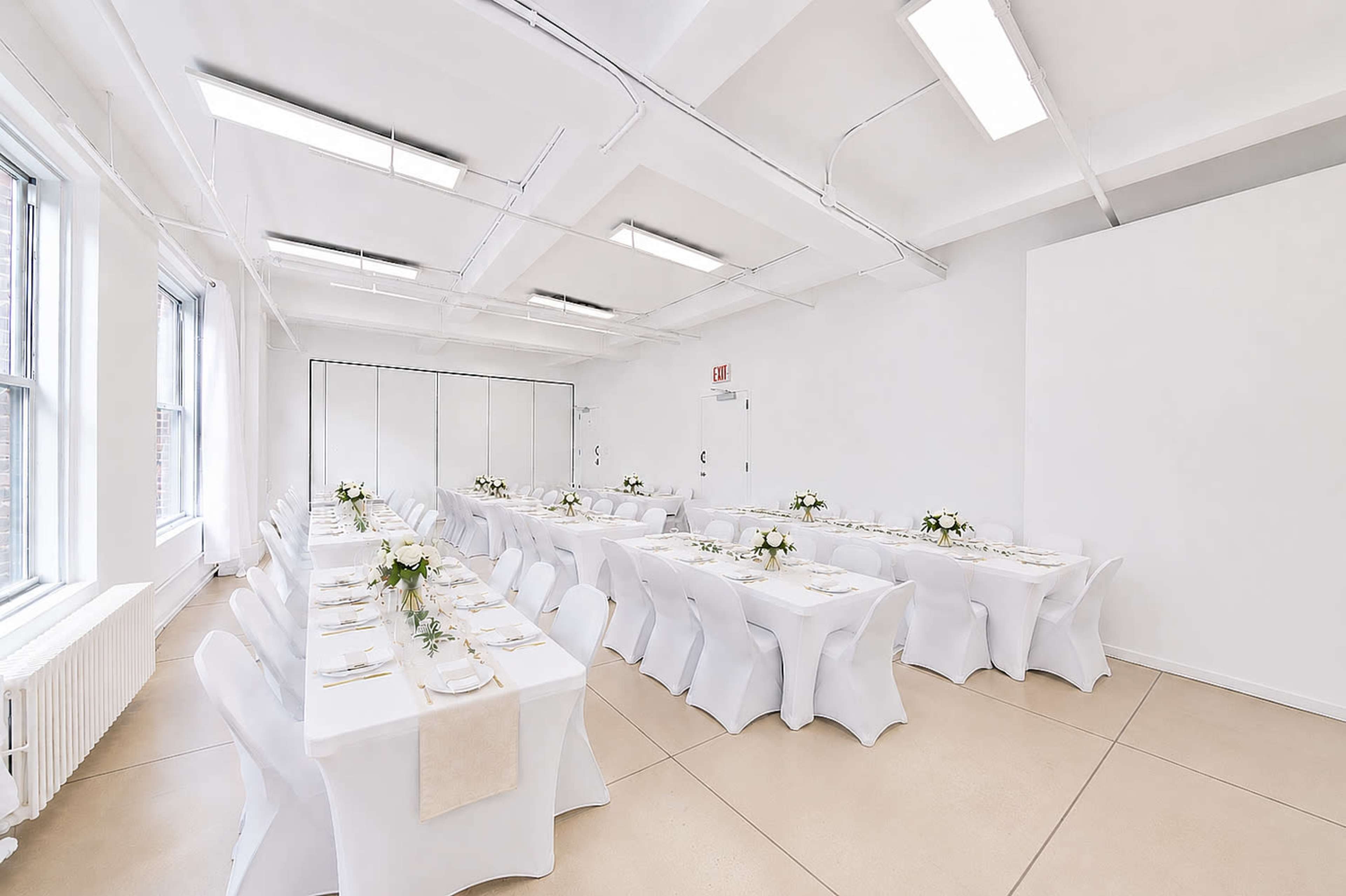 A large, bright room features several tables set with white tablecloths and floral centerpieces for an event.