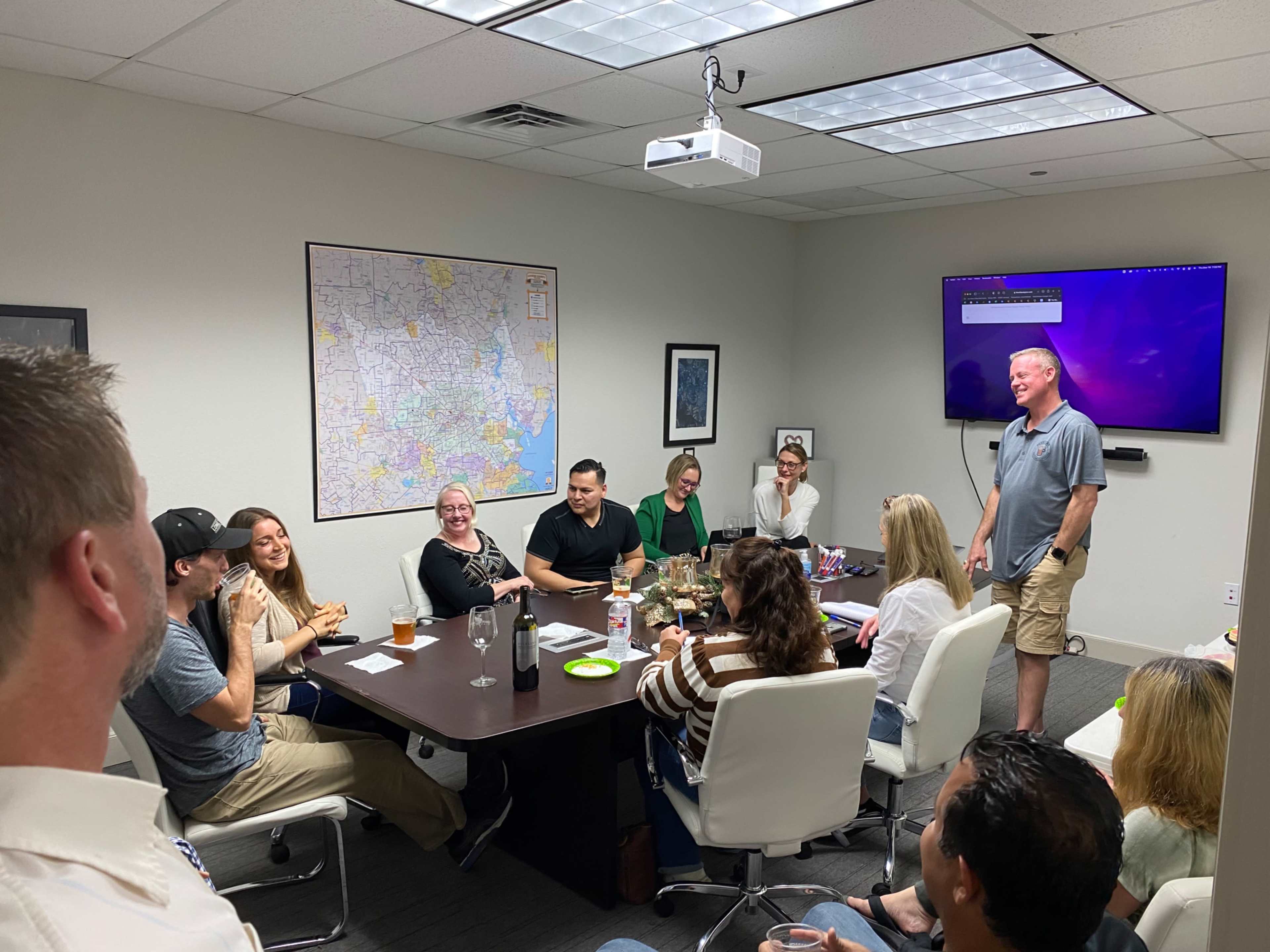 A group of people gathers around a conference table in a meeting room, engaging in conversation while a presentation screen displays information.