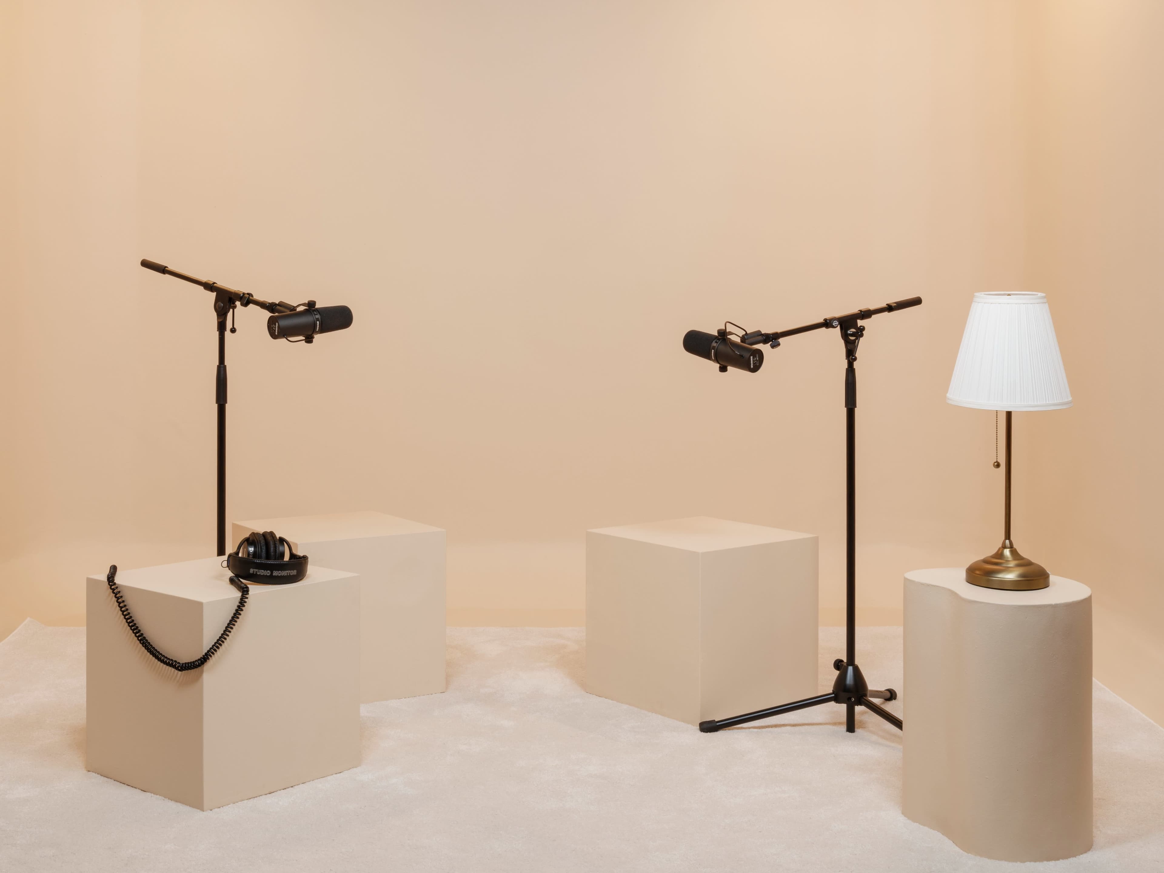 The image shows a minimalistic recording studio setup with three light-colored pedestals, two microphones on stands, a pair of headphones, and a lamp.